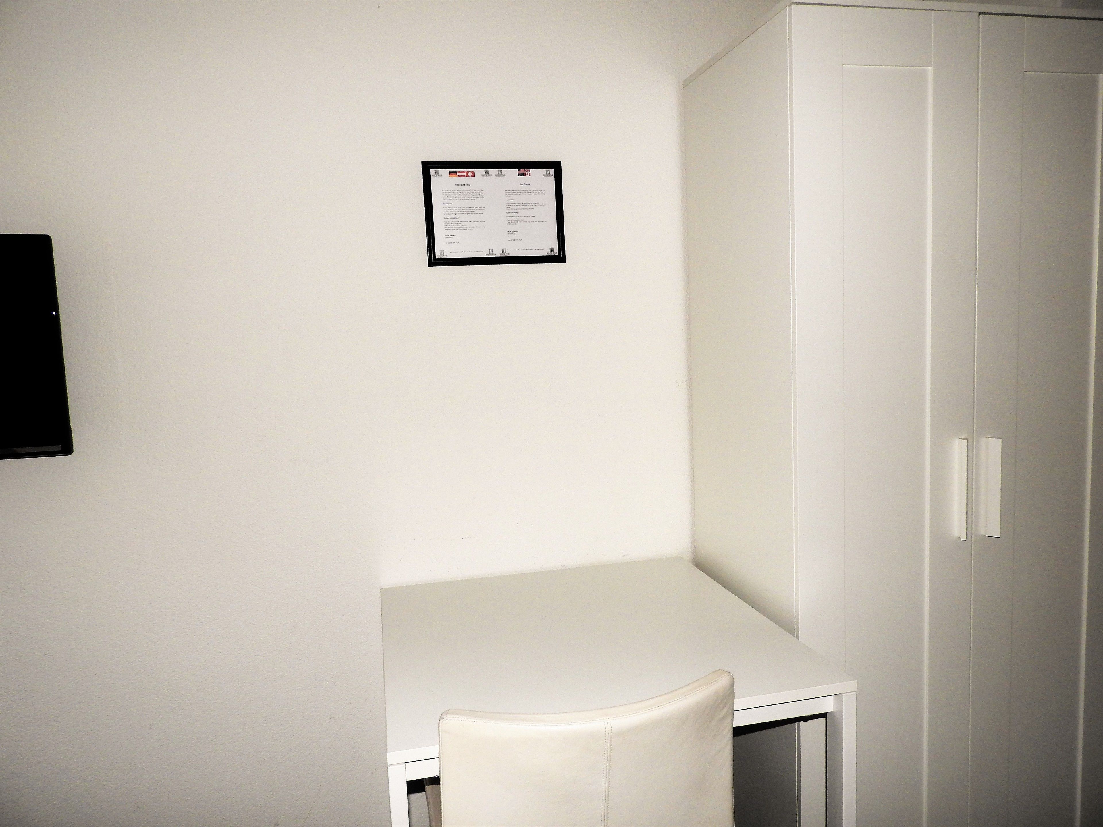 Une petite pièce blanche, avec un bureau et une chaise, une armoire blanche et un avis encadré accroché au mur.