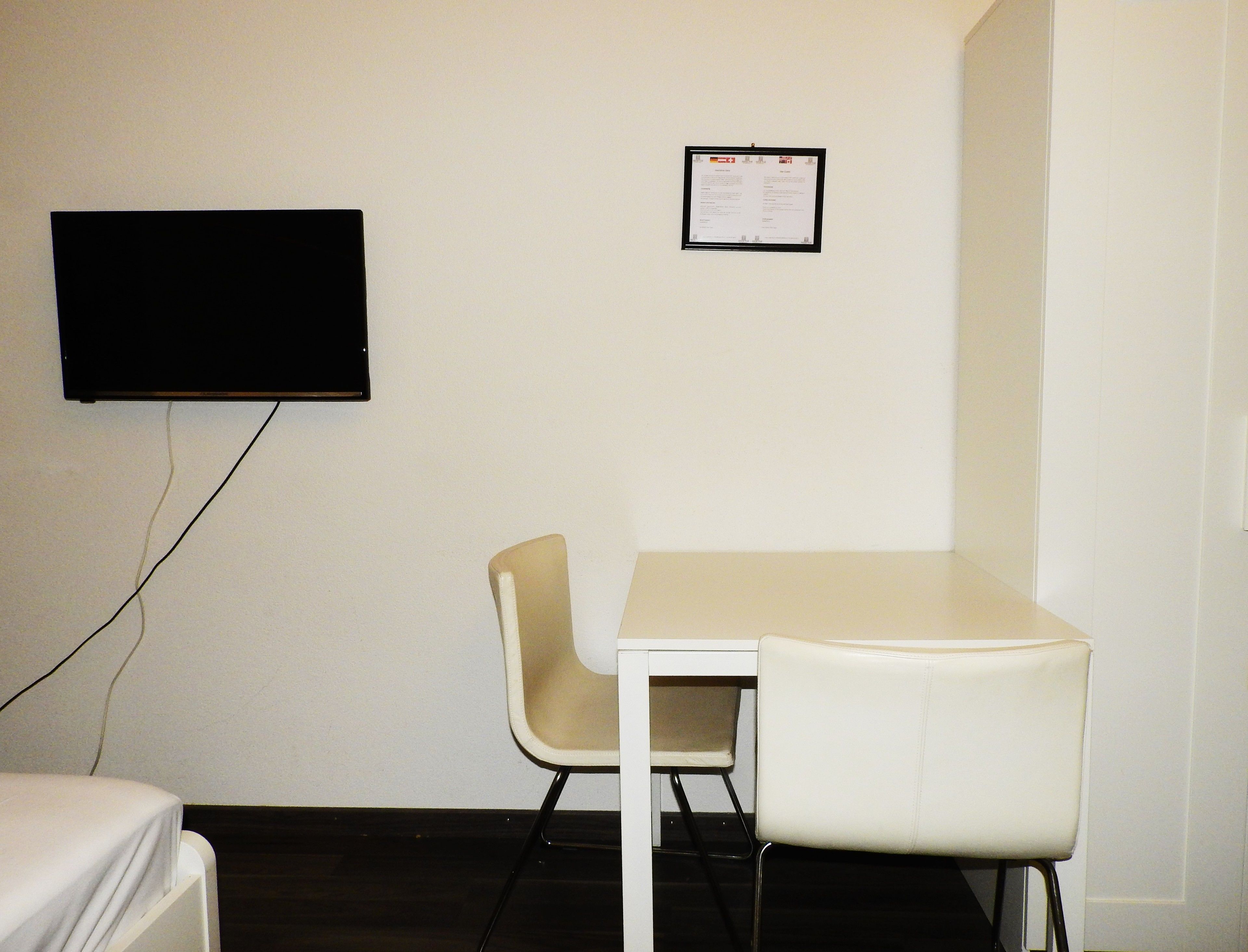 A room with a white table and two chairs, a flat-screen TV mounted on the wall, and a framed notice on the wall.