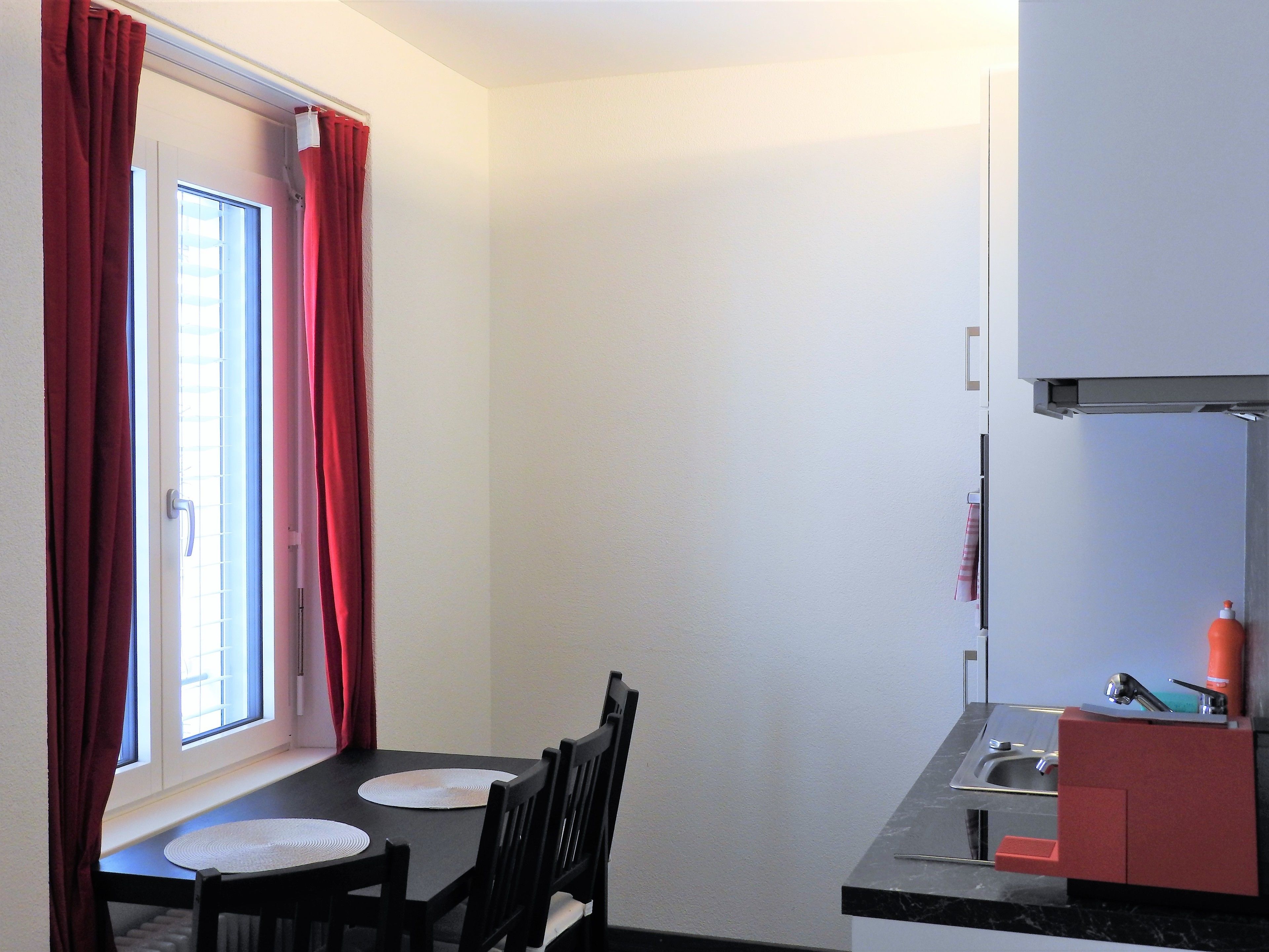Une petite cuisine avec une fenêtre, une table à manger avec deux chaises et un buffet rouge.
