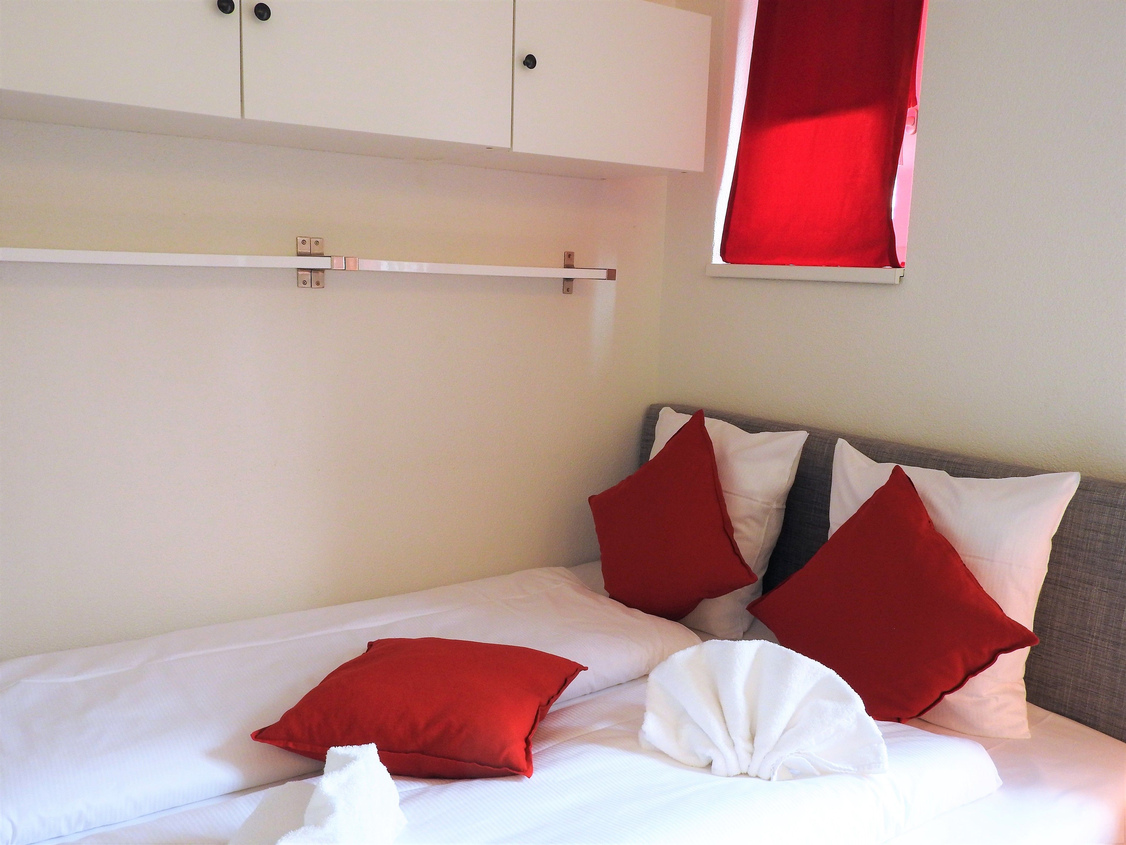 Cama bem arrumada com almofadas brancas e vermelhas, cabeceira cinza e mesa de cabeceira branca.