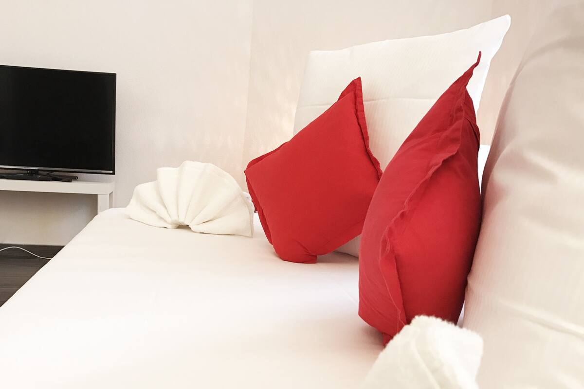 A neatly made bed with white and red pillows, a white blanket, and a flat-screen TV on a white stand.