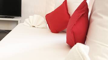 Una cama bien hecha con almohadas blancas y rojas, una colcha blanca y un televisor de pantalla plana sobre un soporte blanco.