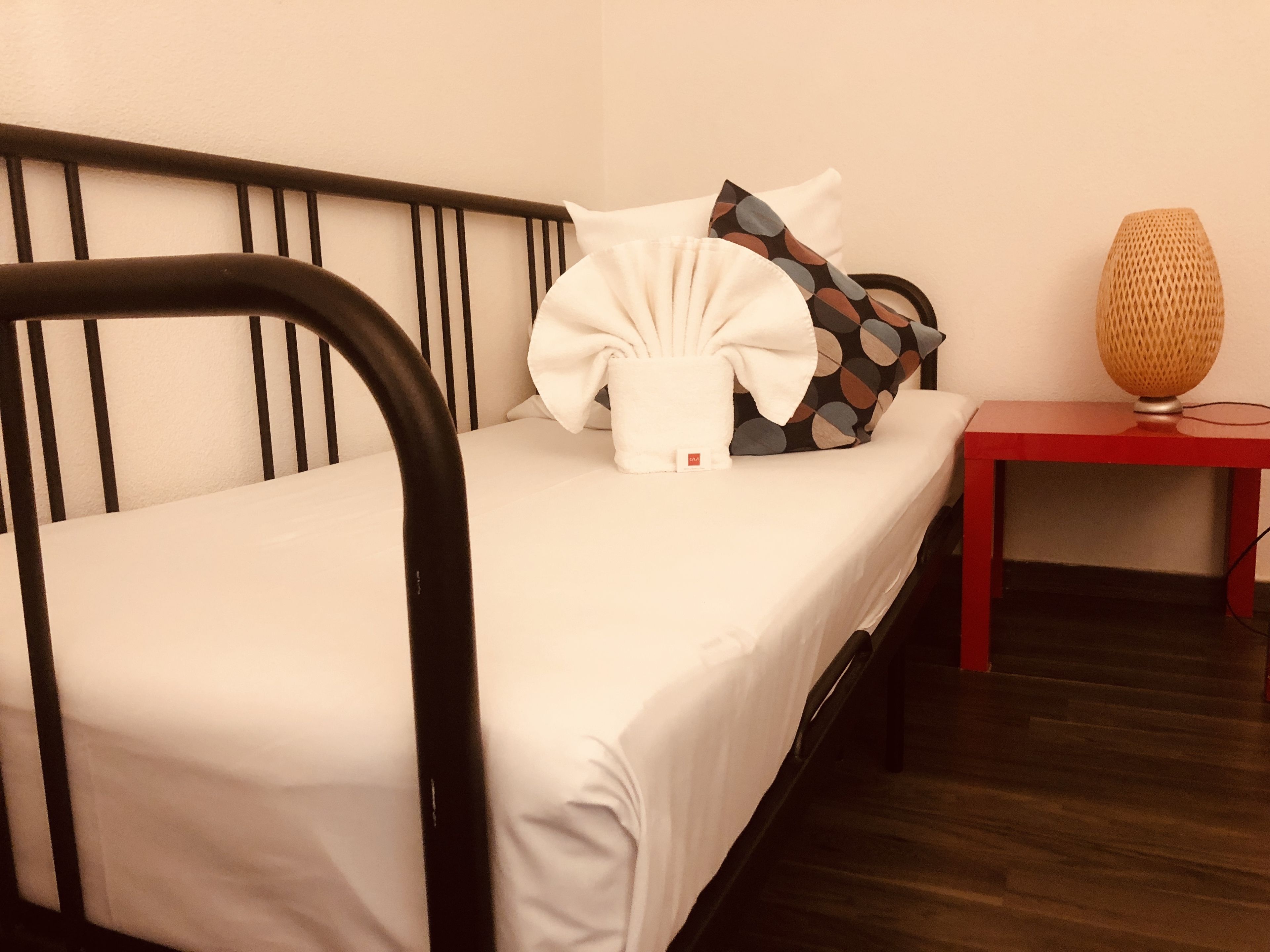 Un lit simple avec un drap blanc, une tête de lit en métal noir, une petite table de chevet rouge avec une lampe à abat-jour en osier, et un coussin à motifs.