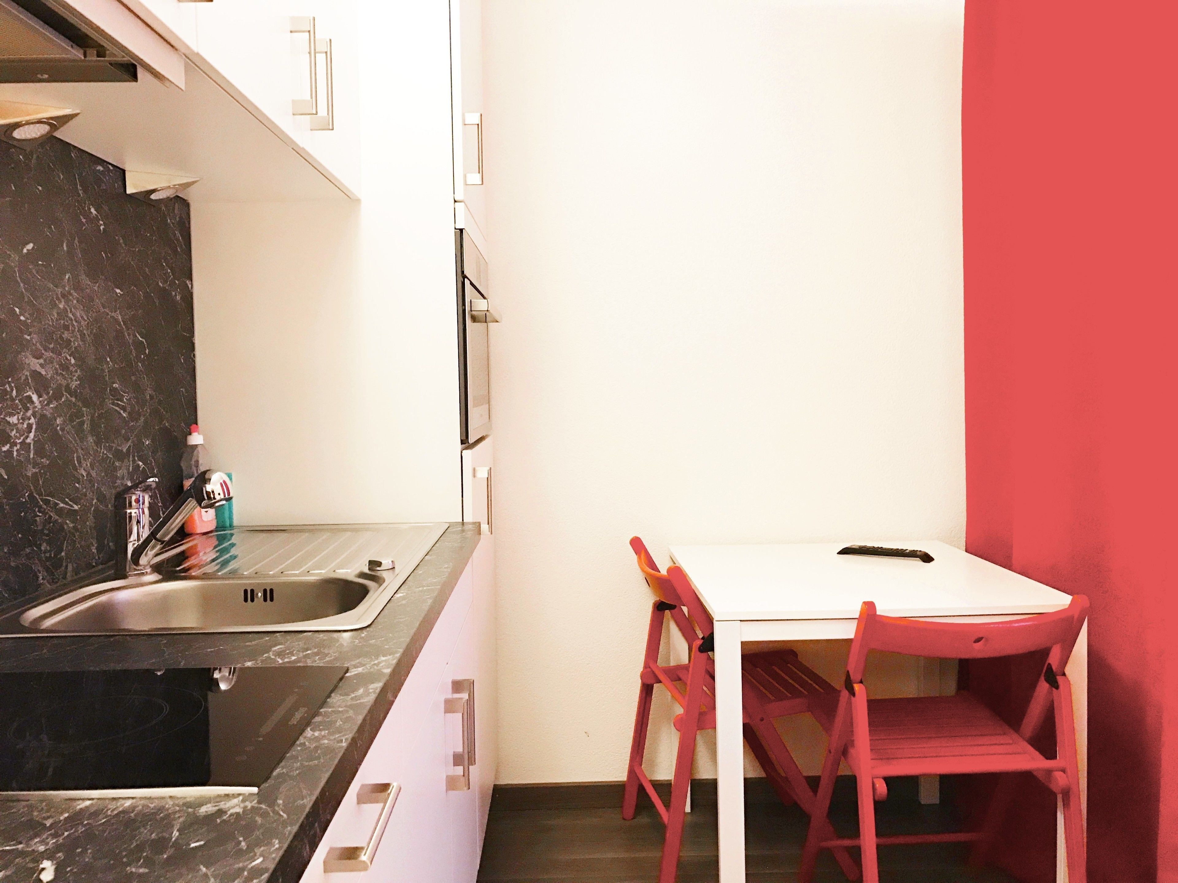 Una piccola cucina con un lavello, un piano di lavoro bianco e una sedia rossa.