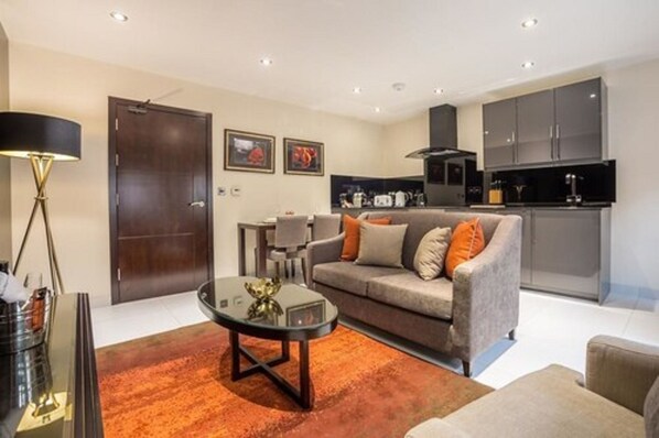 A modern living room with a sofa, a coffee table, and a lamp. There is a kitchen area with cabinets and a stove. - Mansio Suites The Headrow (Leeds)