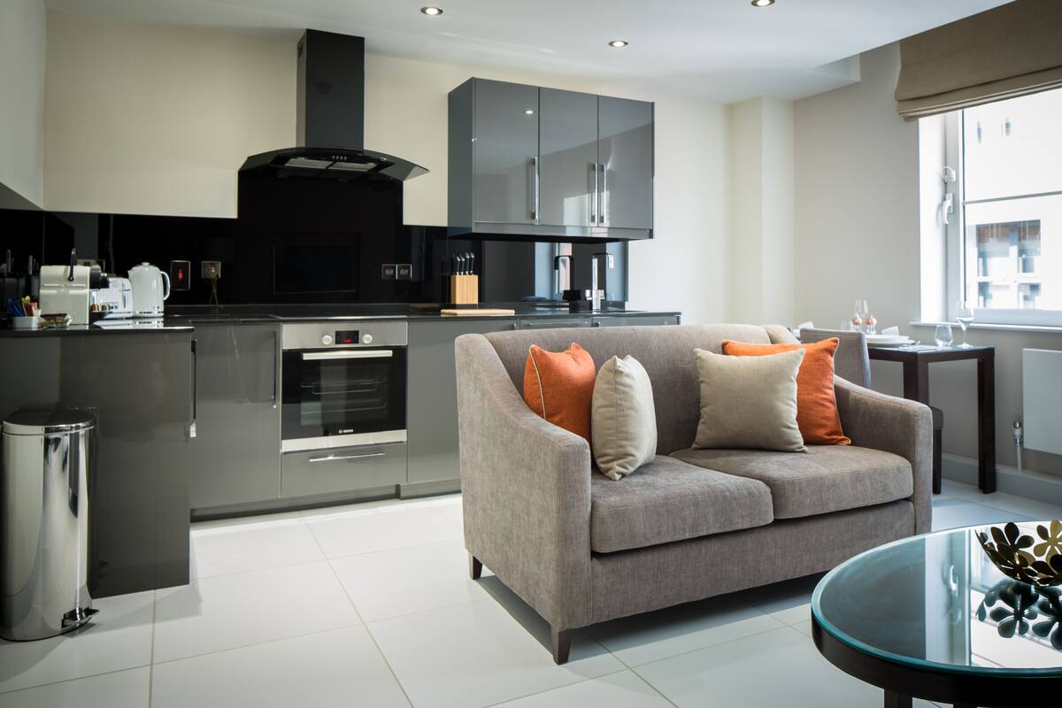 A modern kitchen with grey cabinets, a built-in oven, and a grey sofa with orange cushions.