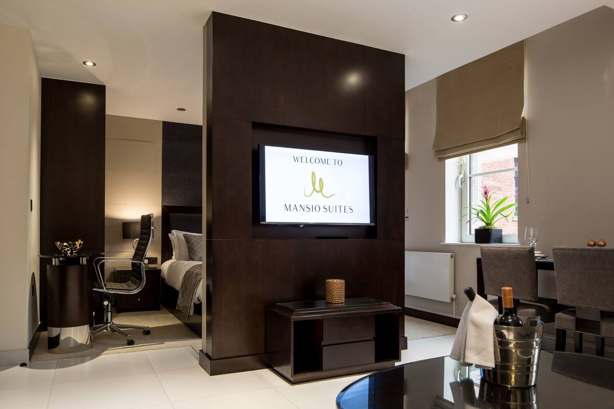 A modern hotel room with a bed, a desk, a chair, a TV mounted on the wall, and a window with a plant.