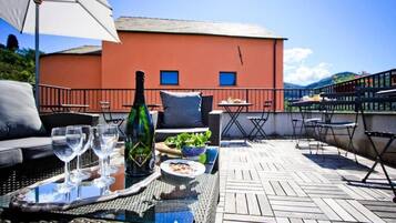 A terrace with outdoor furniture, a table with a bottle and glasses, and a view of a red building.