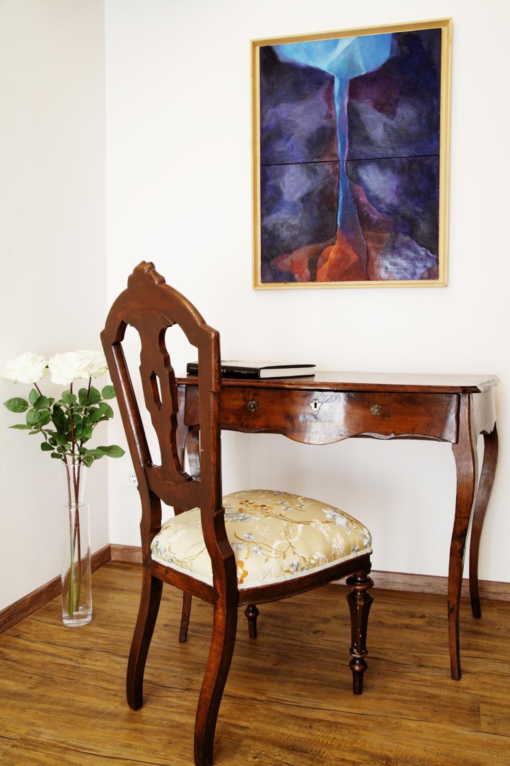 A wooden desk with a chair, a vase with flowers, and a painting on the wall.