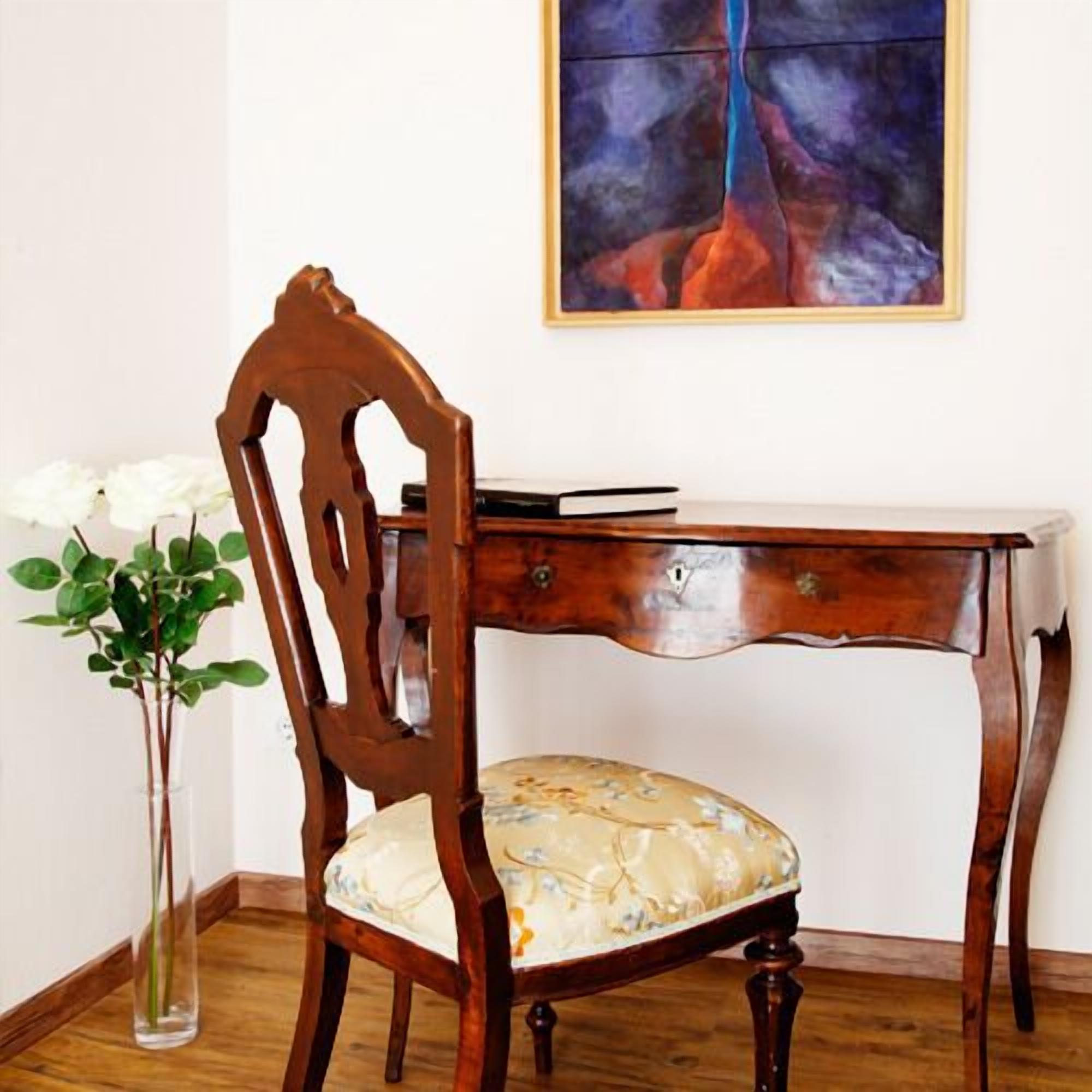 A wooden desk with a cushioned chair, a vase with white flowers, and a painting on the wall.