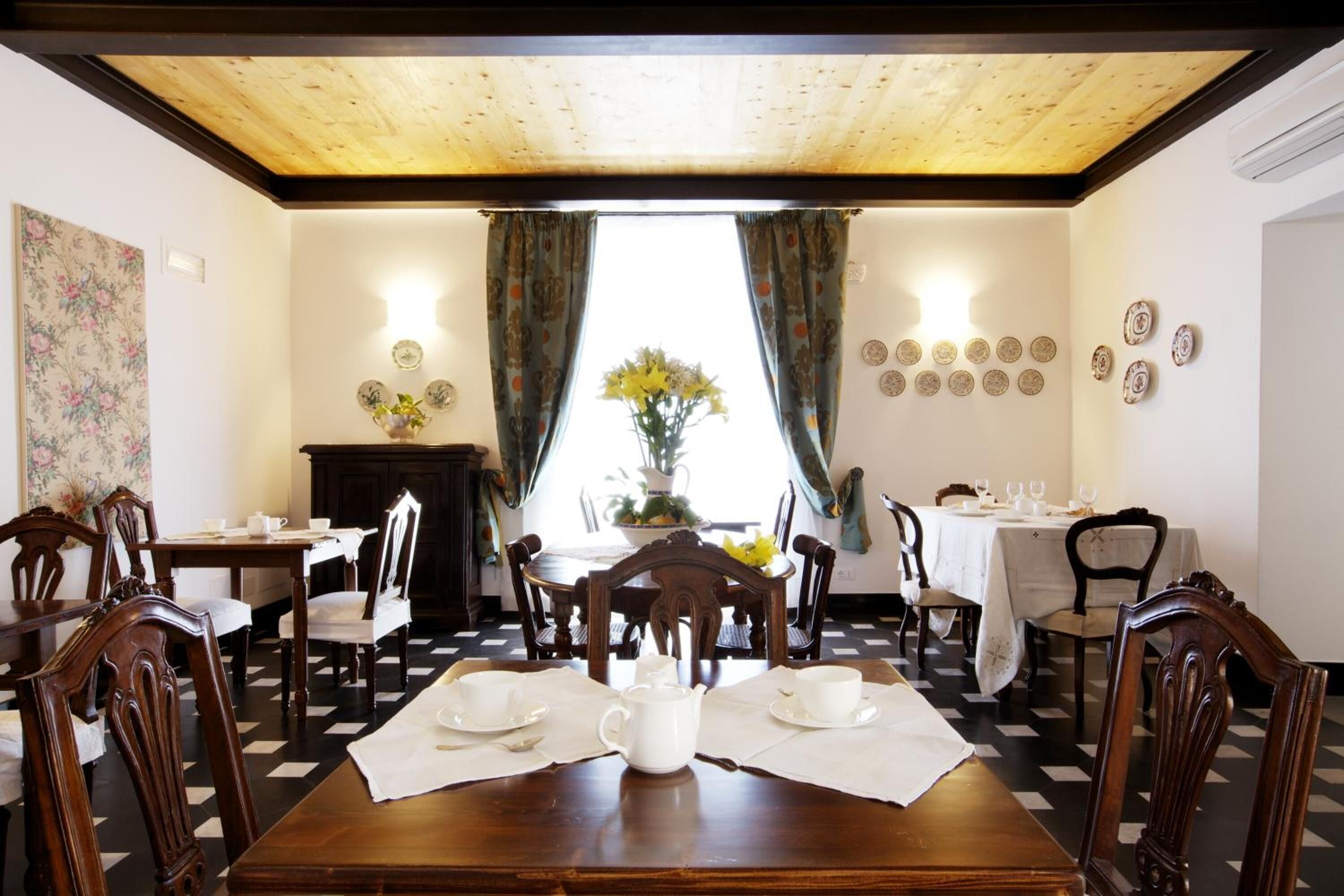 A dining room with a wooden table set for four, surrounded by chairs. There are two windows with curtains, a wall with decorative plates, and a floral painting on the wall.