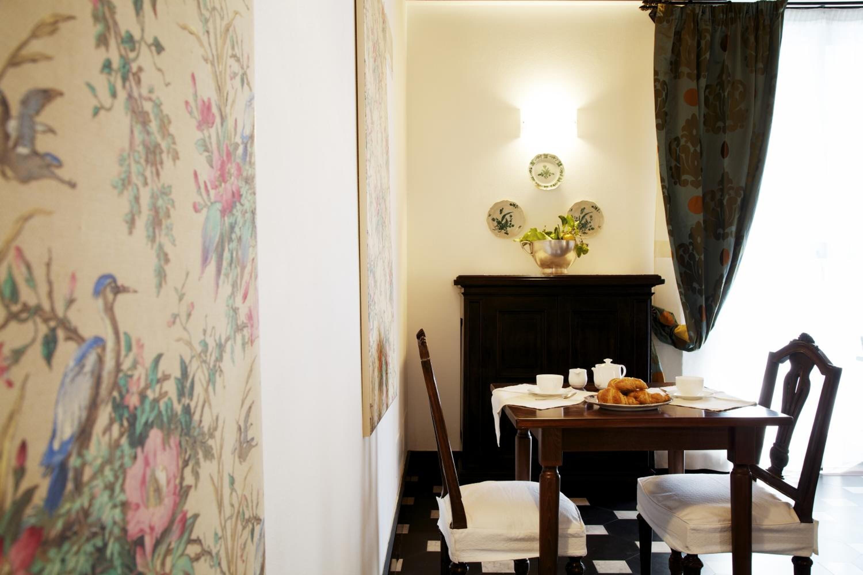 Un coin repas avec une table dressée pour deux personnes, un vase rempli de fleurs et un mur orné d’un tableau représentant des motifs floraux.