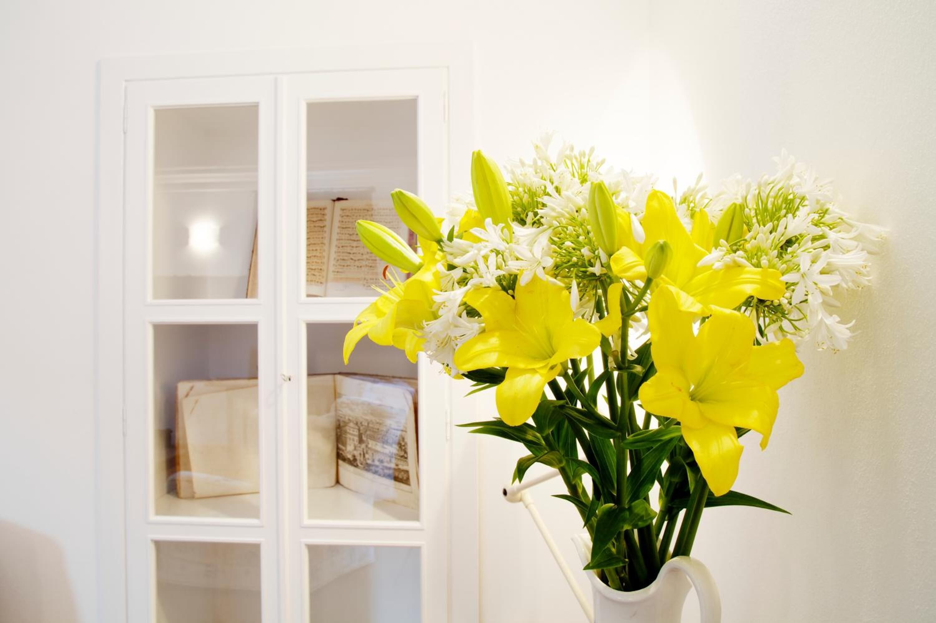 A white cabinet with glass doors containing books and a white vase with yellow and white flowers.