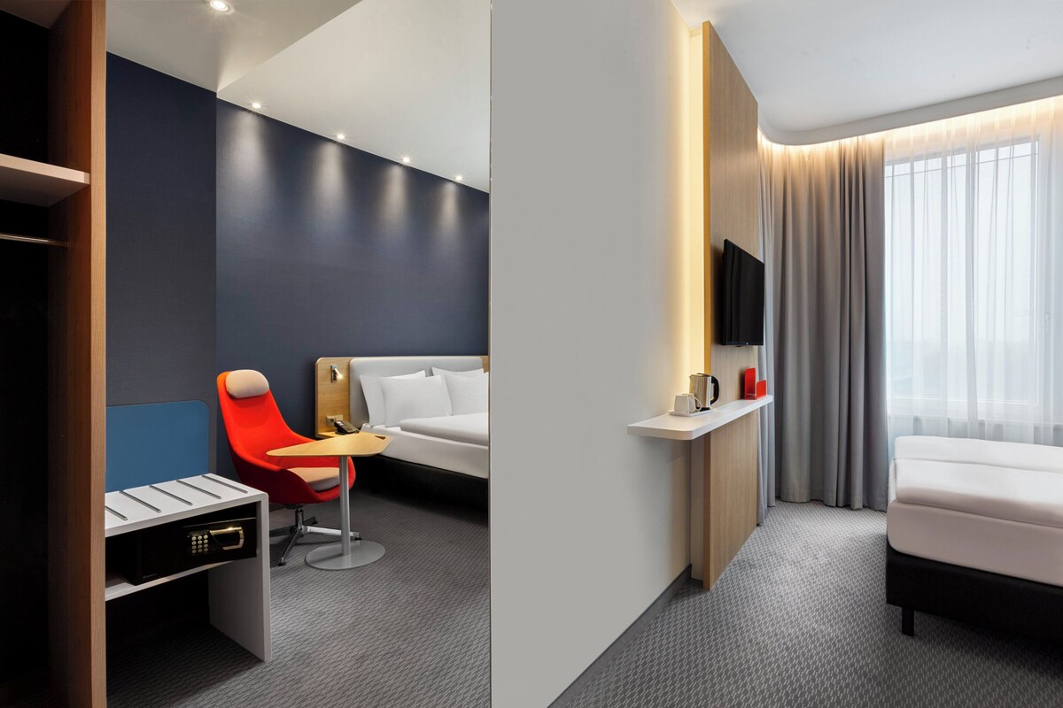 A modern hotel room with a bed, a desk with a chair, a TV, and a window with curtains.