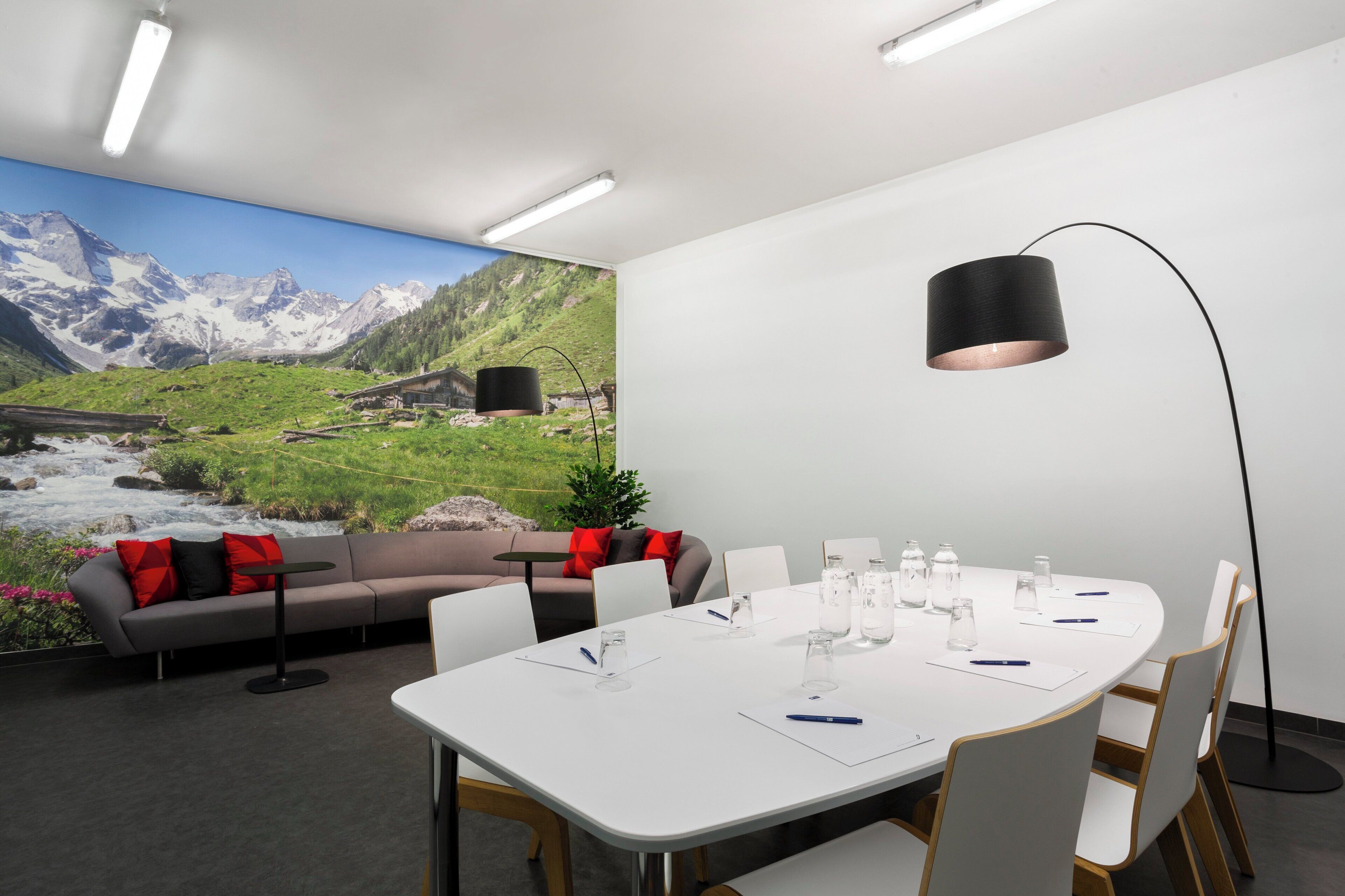 Een vergaderruimte met een witte tafel, stoelen, een bank met rode kussens, een vloerlamp en een muurschildering van een berglandschap.