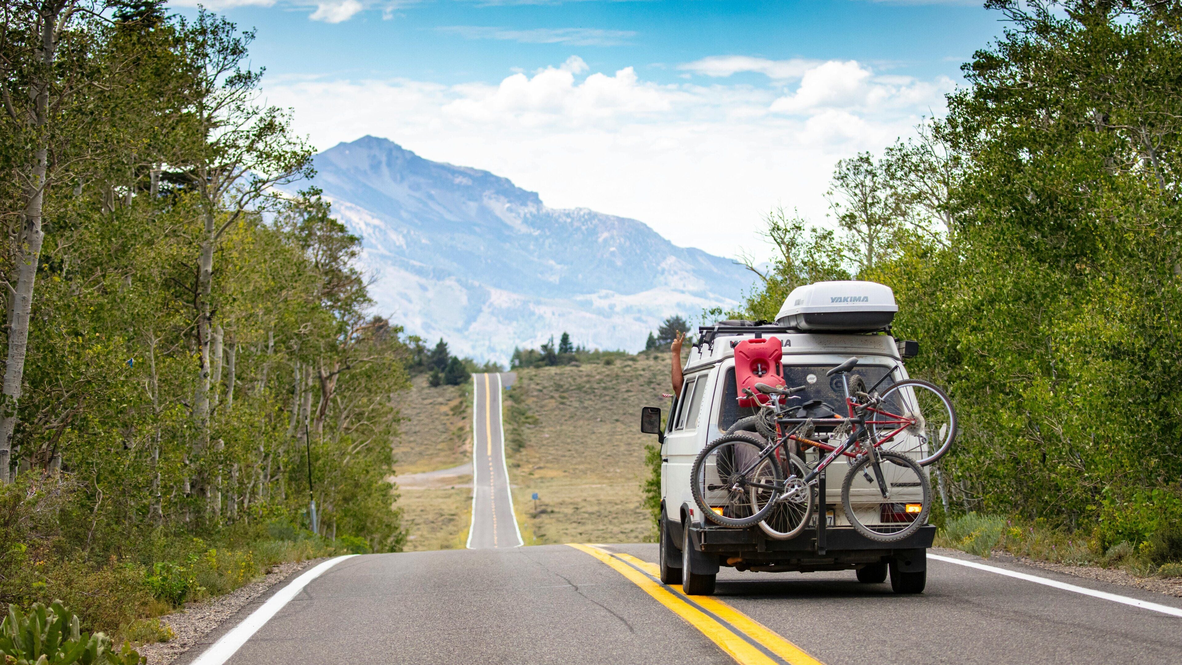 A vehicle with bicycles mounted on the back, traveling on a road flanked by trees and mountains in the background.