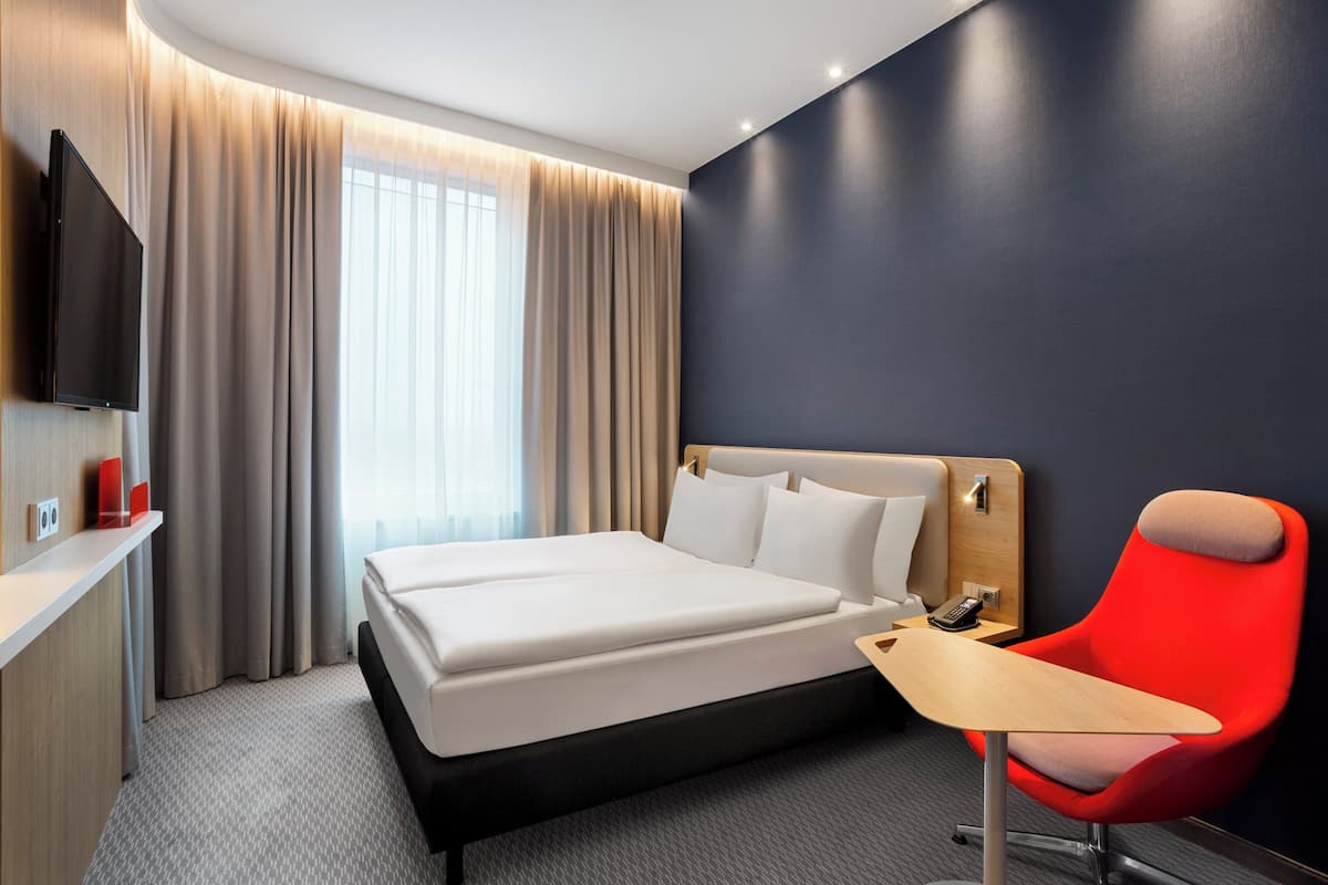 A hotel room with a bed, a TV mounted on the wall, a desk, and a red chair.