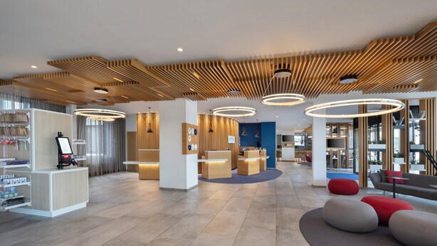 A modern hotel lobby with a reception desk, seating areas, and a large window offering a view of the outdoors. - Holiday Inn Express Munich City West by IHG (Munich)