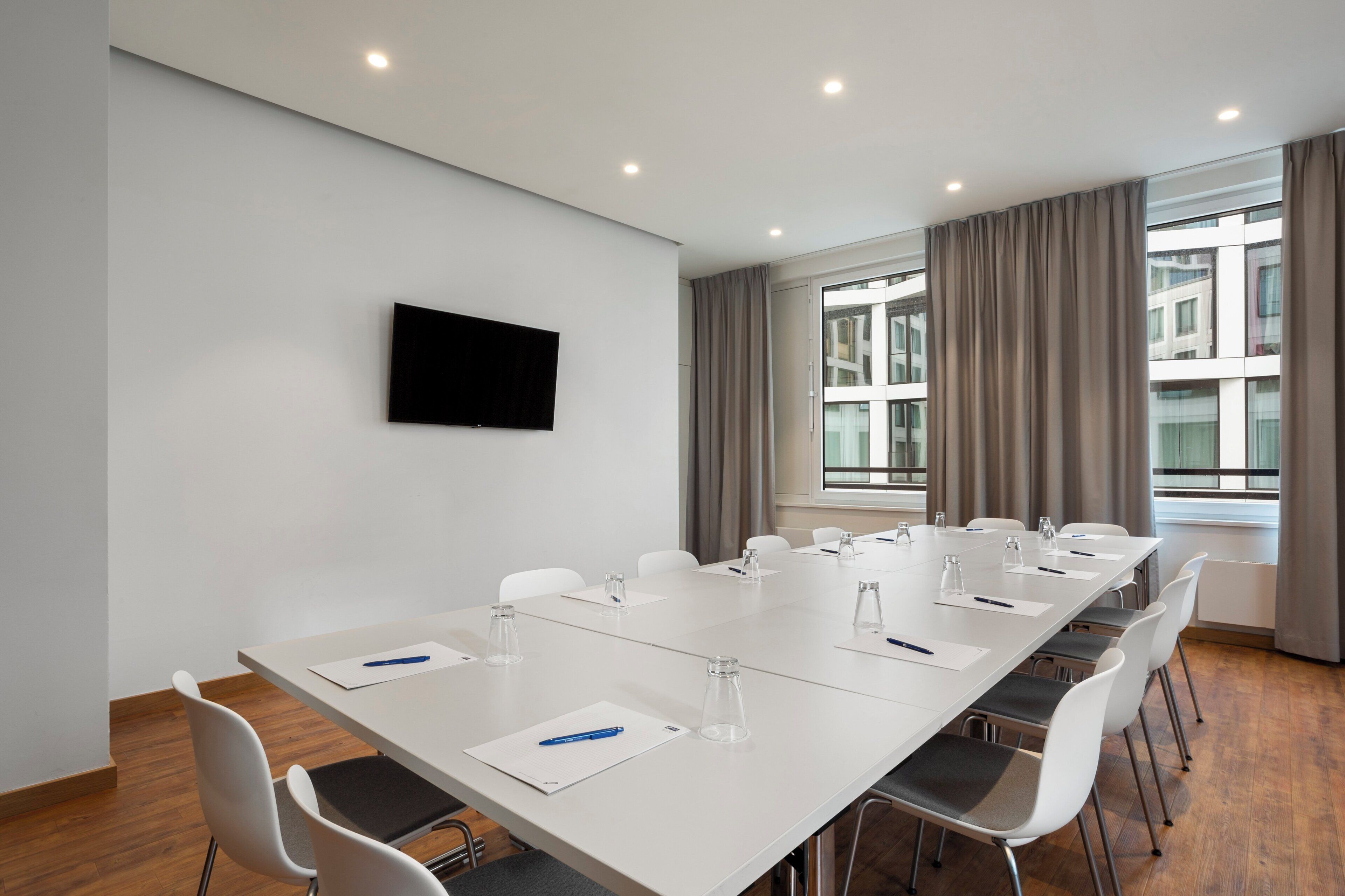 A conference room with a long table, white chairs, a flat-screen TV on the wall, and large windows with curtains.