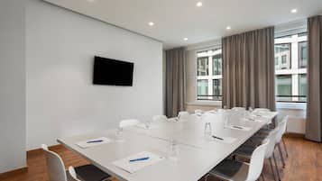 A conference room with a long table, white chairs, a flat-screen TV on the wall, and large windows with curtains.