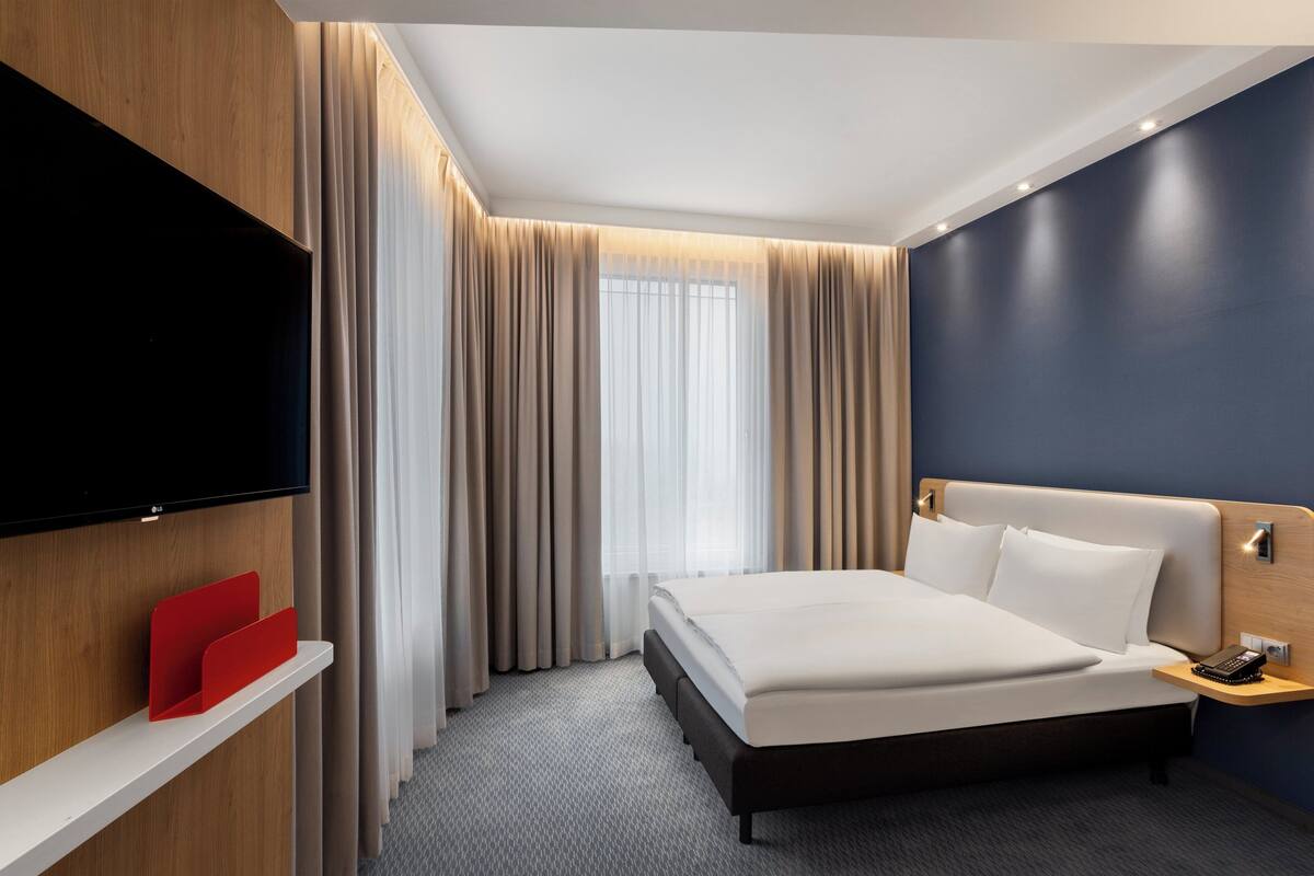 A modern hotel room with a large bed, a flat-screen TV mounted on the wall, a bedside table with a phone, and a window with sheer curtains.