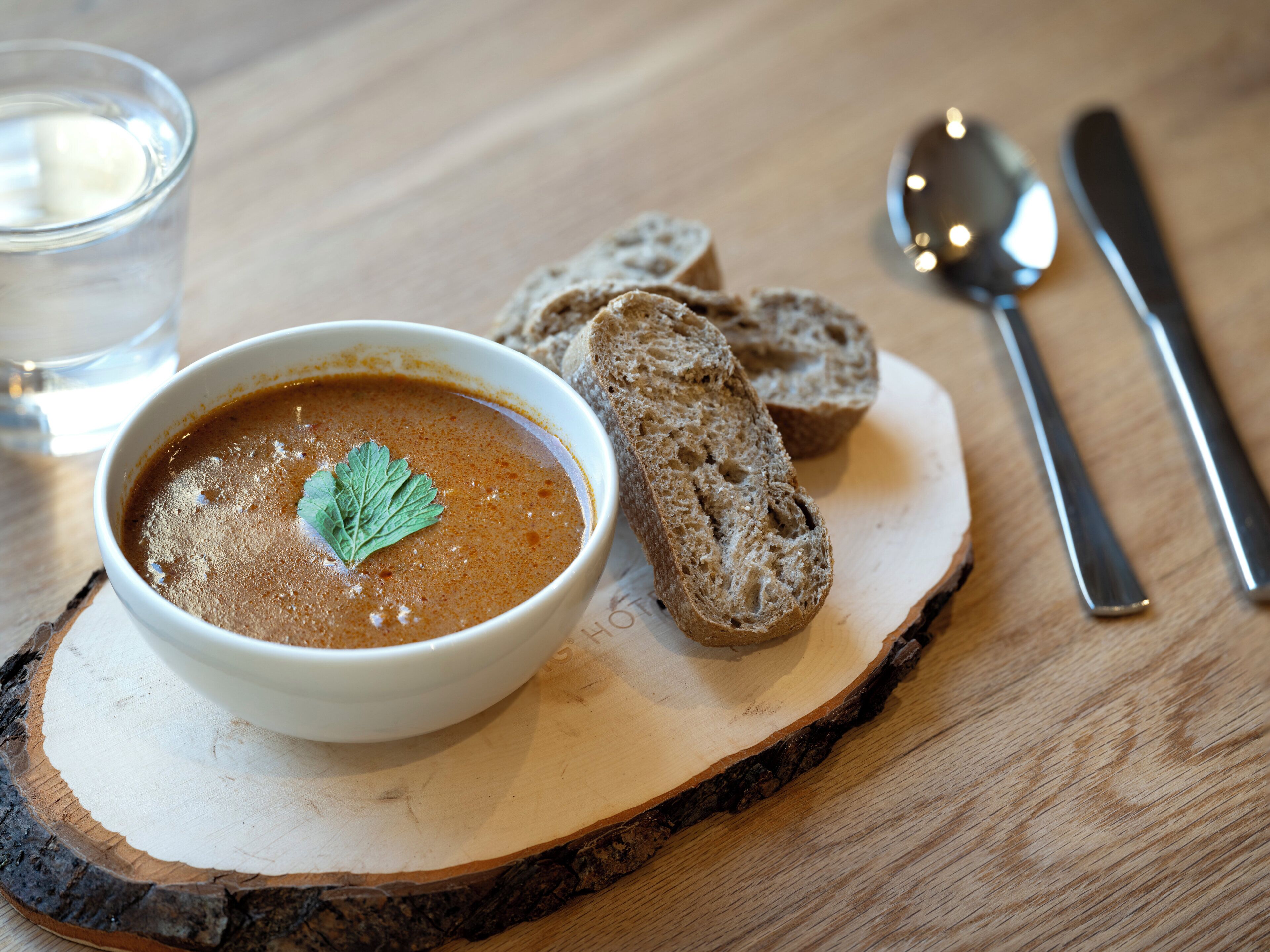 A bowl of soup with a garnish on a wooden board, accompanied by slices of bread and a spoon.