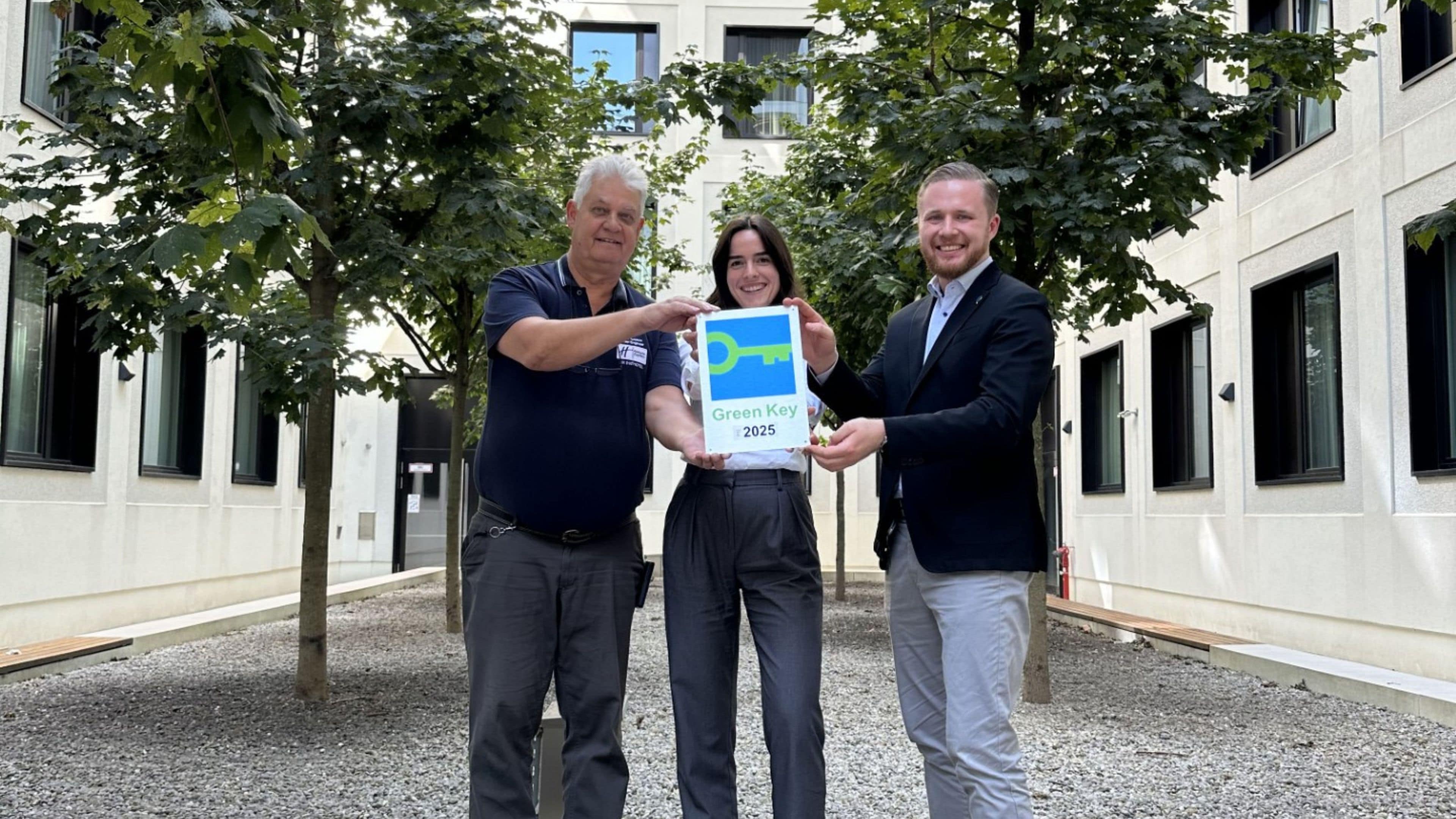 Drei Personen halten ein Schild mit der Aufschrift 'Green Key 2025' vor einem Gebäude mit Innenhof.