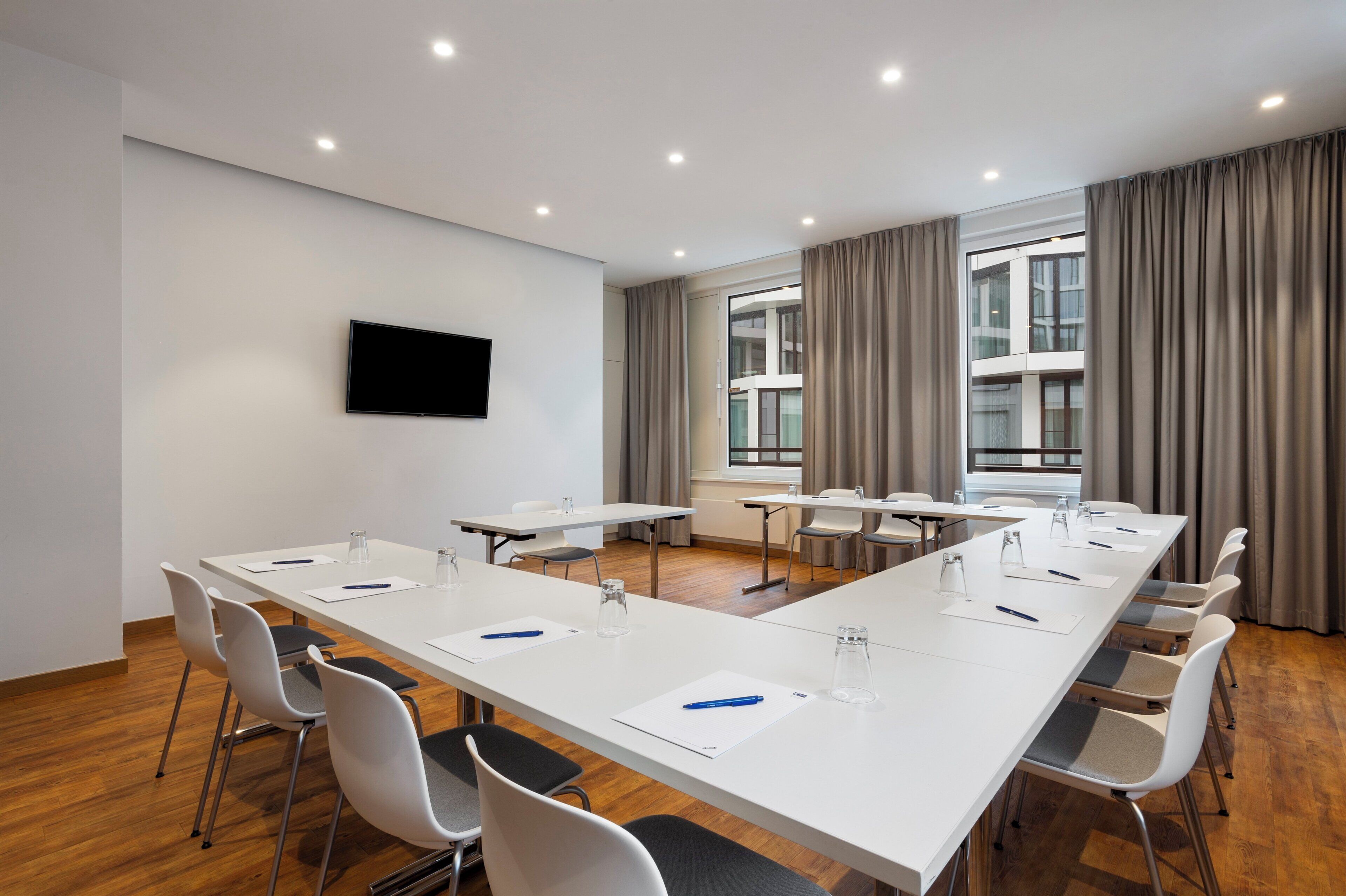 A conference room with a long table, chairs, a flat-screen TV, and large windows with curtains.