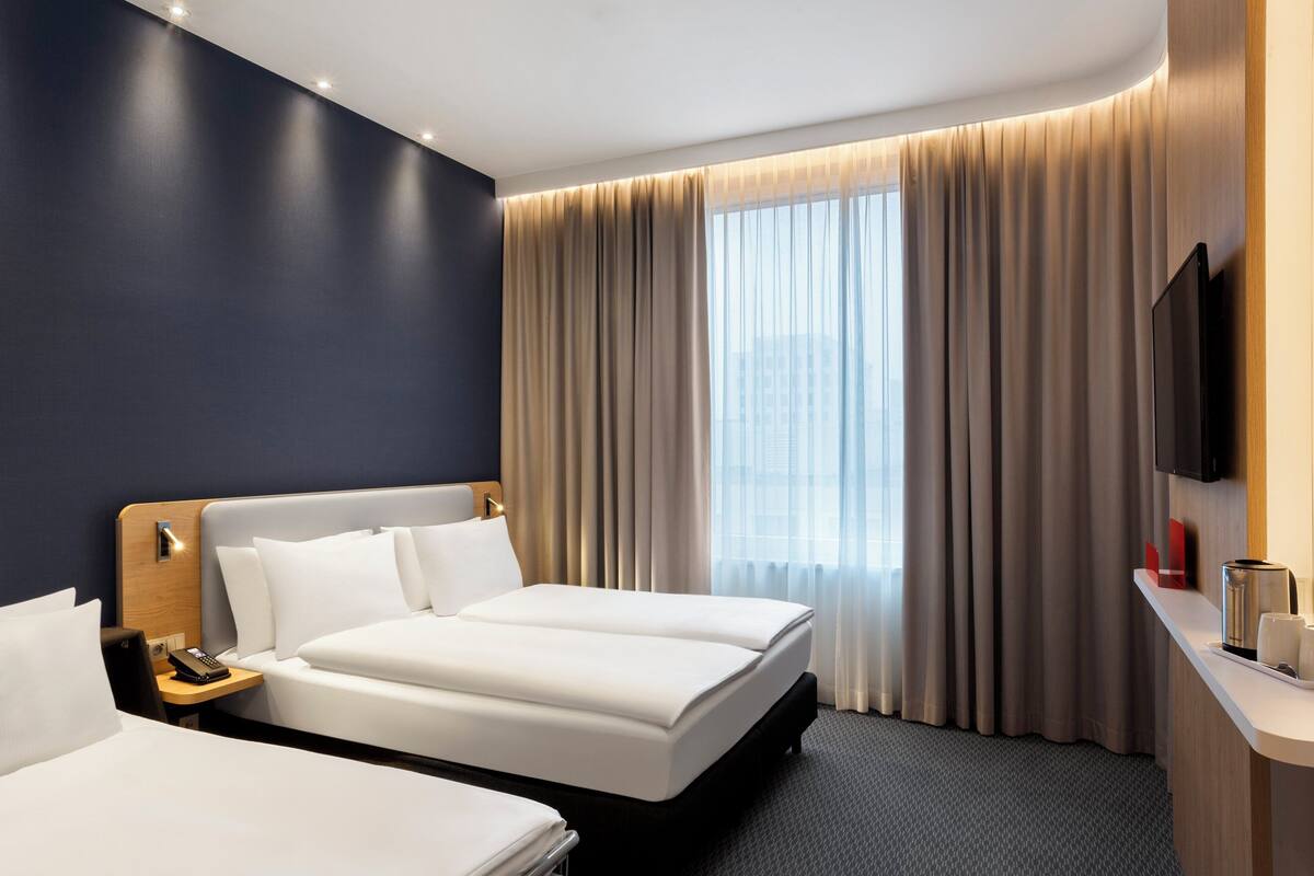 A hotel room with two beds, a flat-screen TV, a bedside table with a phone, and a large window with curtains.