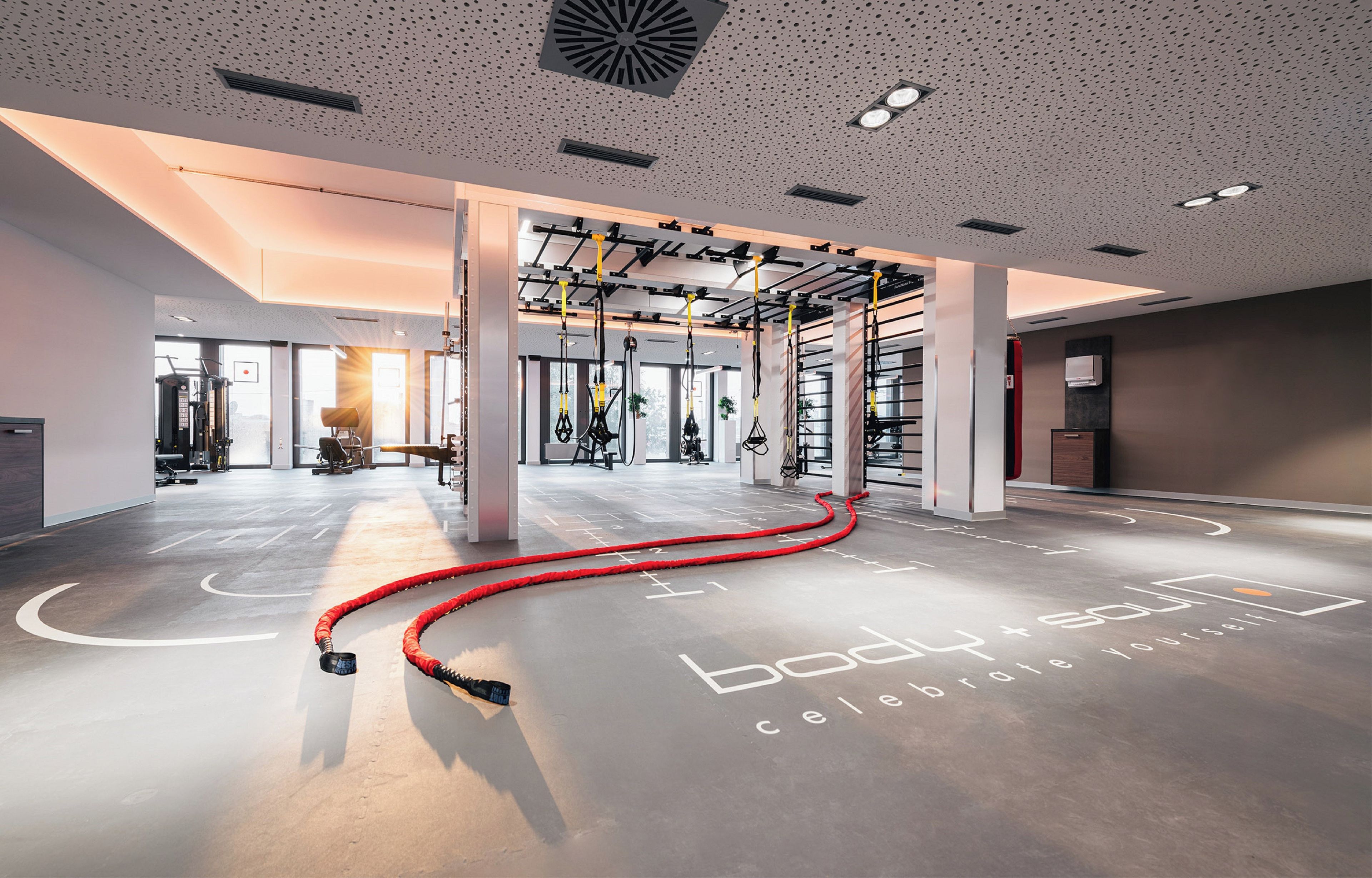 Ein geräumiger Fitnessraum mit hängenden Trainingsgeräten, einem roten Schlauch auf dem Boden und motivierenden Text.