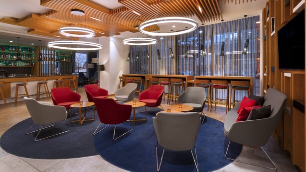 Un espace de détente moderne avec des panneaux de plafond en bois, des luminaires suspendus de forme circulaire et divers types de sièges, notamment des chaises rouges et grises.