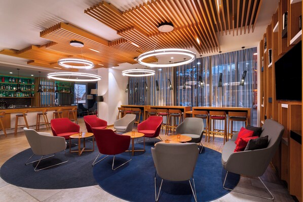 A modern lounge area with wooden ceiling panels, circular pendant lights, and a variety of seating options including red and grey chairs. - Holiday Inn Express Munich City West by IHG (Munich)