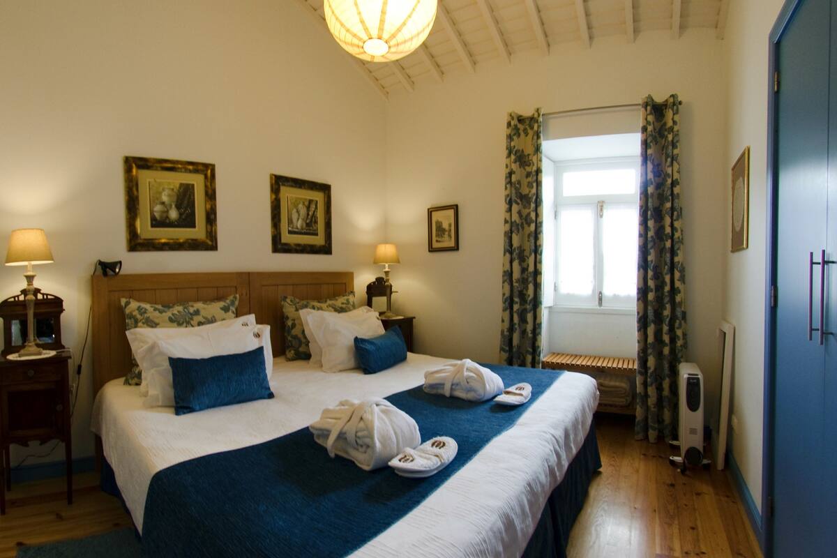 Ein Hotelzimmer mit einem großen Bett, zwei Nachttischen, einem Fenster mit Vorhängen und gerahmten Bildern an der Wand.