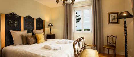 A well-appointed bedroom with a large bed, a wooden headboard, a chandelier, a window with curtains, a chair, and a floor lamp.