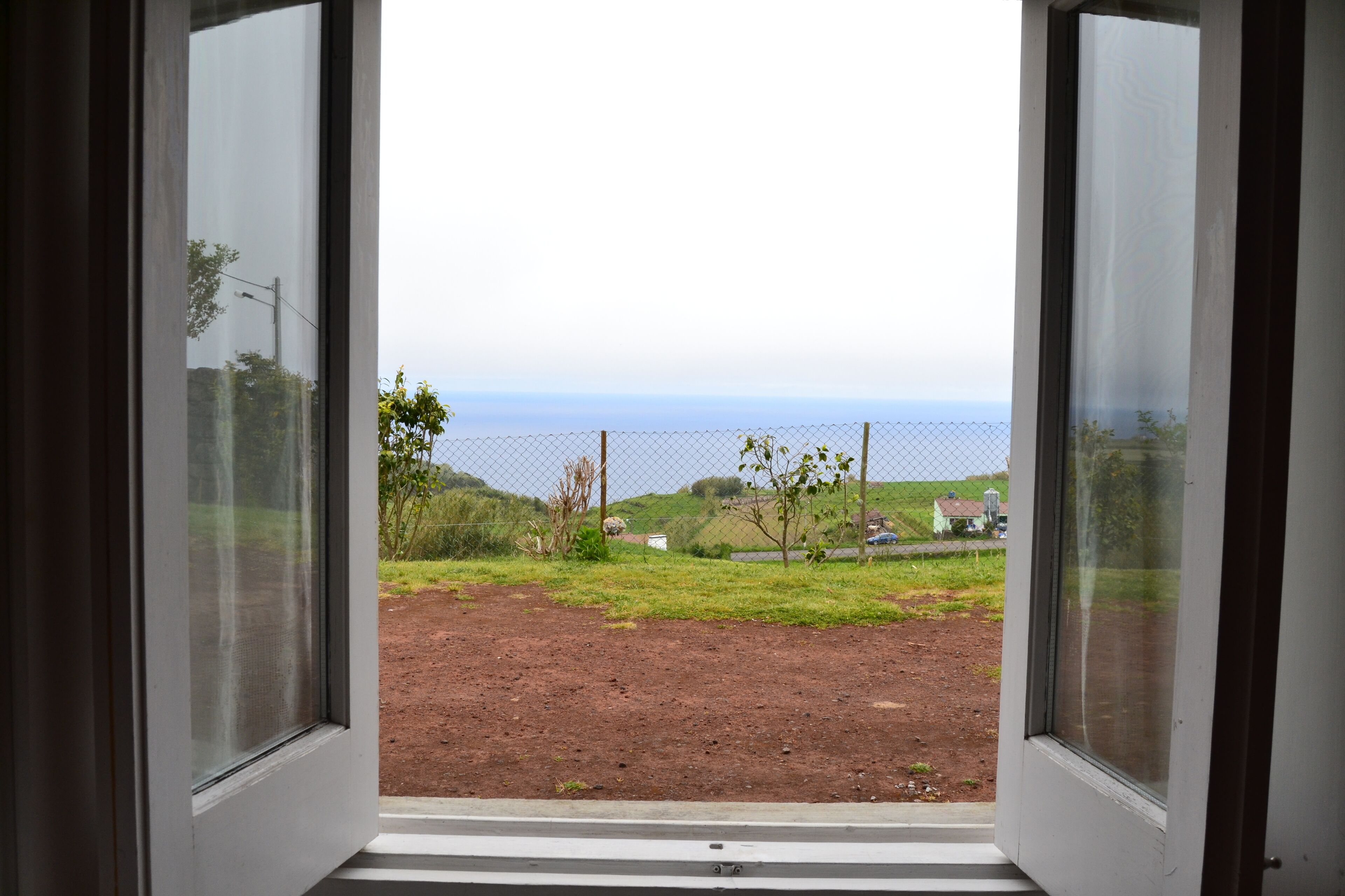 An open window with a view of a grassy area, a fence, and a distant body of water.