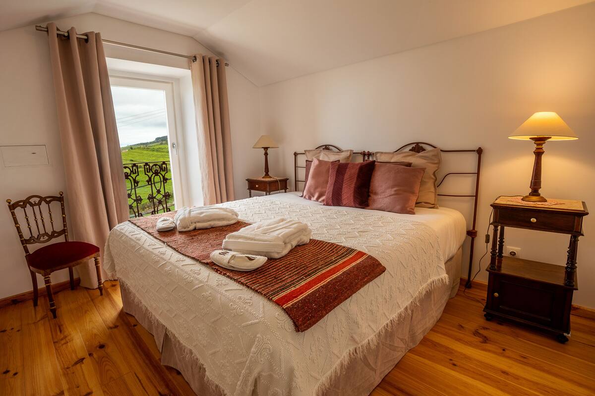 Ein Schlafzimmer mit einem großen Bett, einem Holzstuhl, einem Nachttisch mit Lampe und Blick ins Freie.