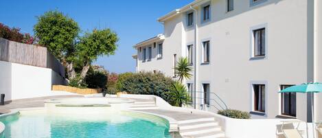 A swimming pool with steps leading into it, surrounded by a white building with multiple windows and a blue sky.