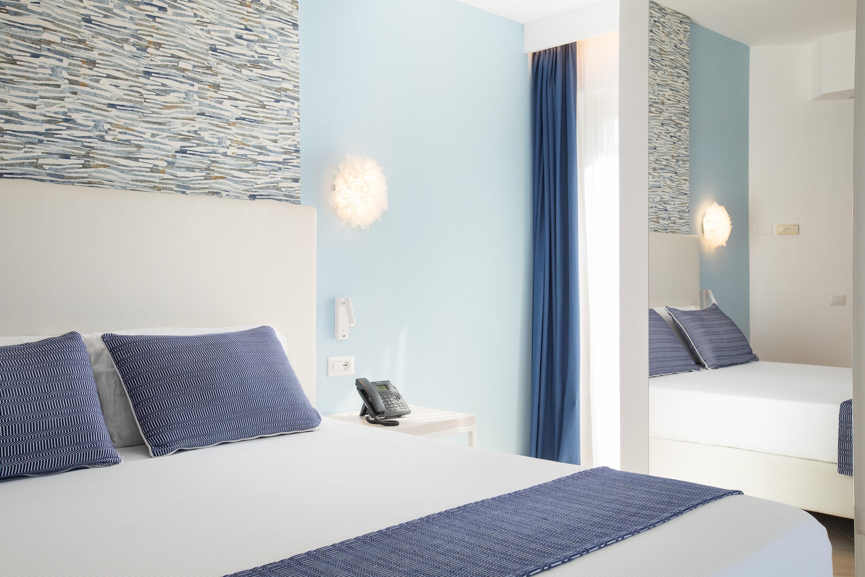 A hotel room with a bed, a bedside table with a phone, and a mirror reflecting the room.