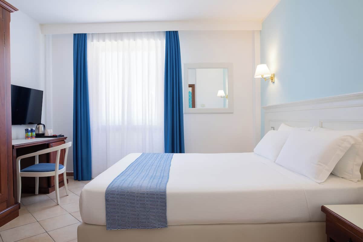Una camera d'albergo con un letto, una scrivania con una sedia, una televisione e una finestra con tende.
