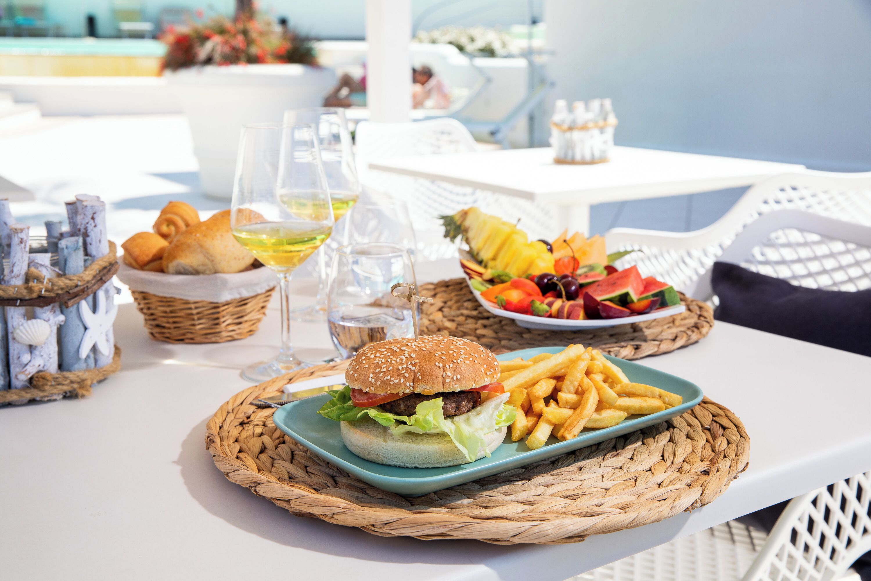 A table with a burger, fries, and a fruit bowl. There are also wine glasses and a basket of bread.