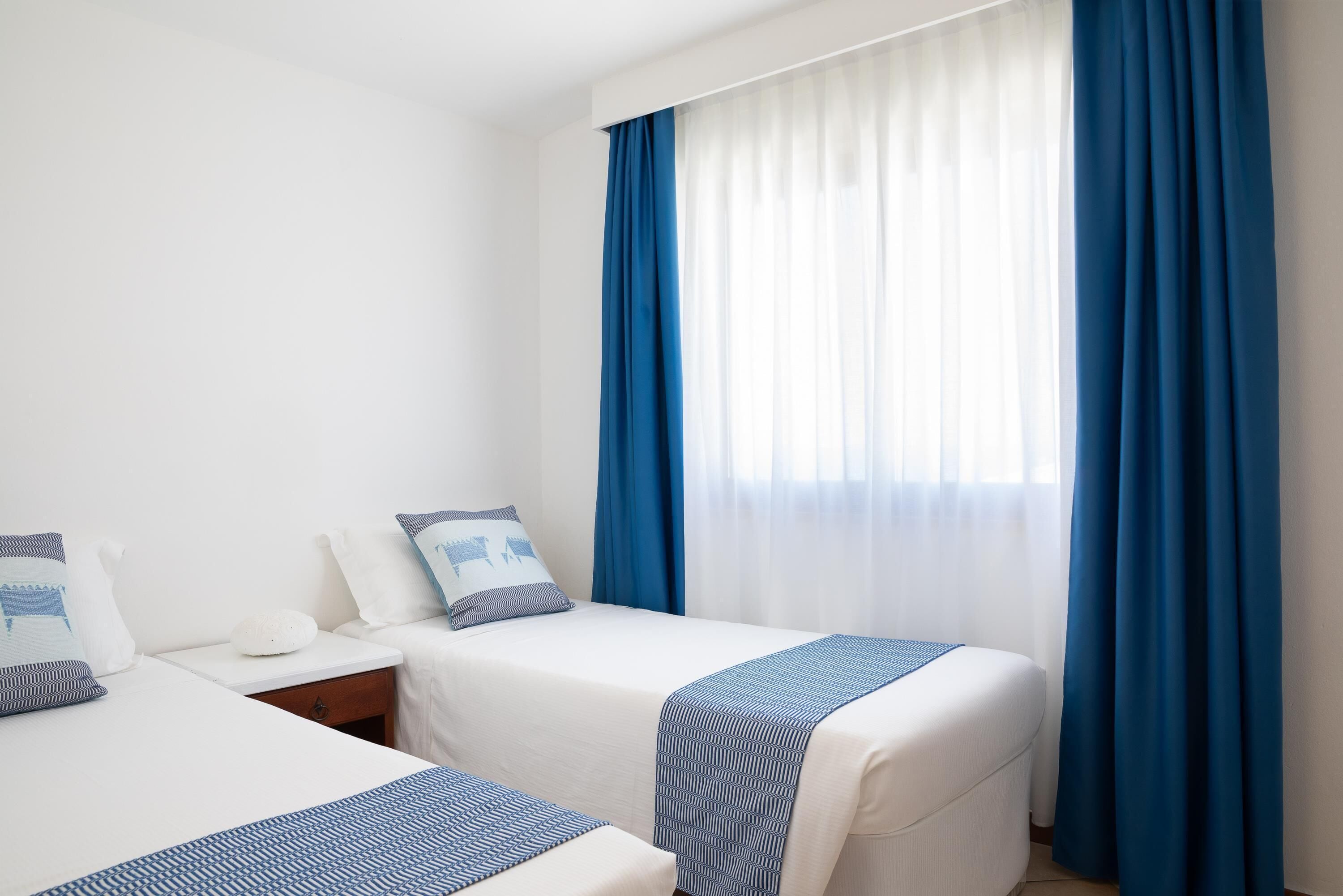 Une chambre d’hôtel avec deux lits, une table de chevet et une fenêtre avec des rideaux bleus.