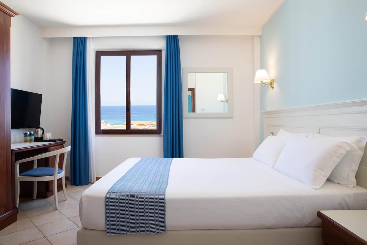 Una camera d'albergo con un letto, una scrivania, una sedia, una televisione e una finestra vista spiaggia.