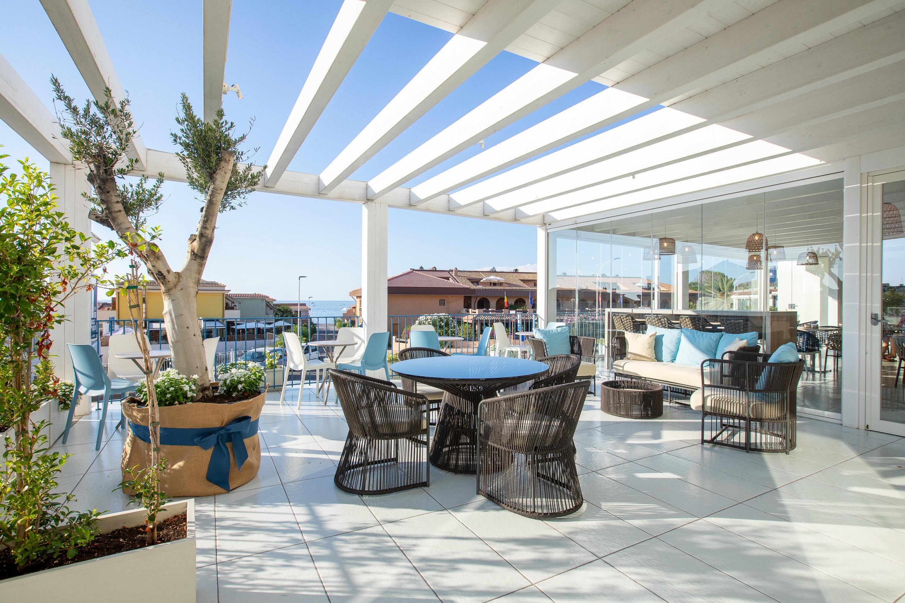 An outdoor patio with a round table and wicker chairs, surrounded by potted plants and a white pergola.