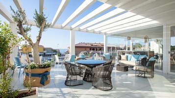 An outdoor patio with a round table and wicker chairs, surrounded by potted plants and a white pergola.