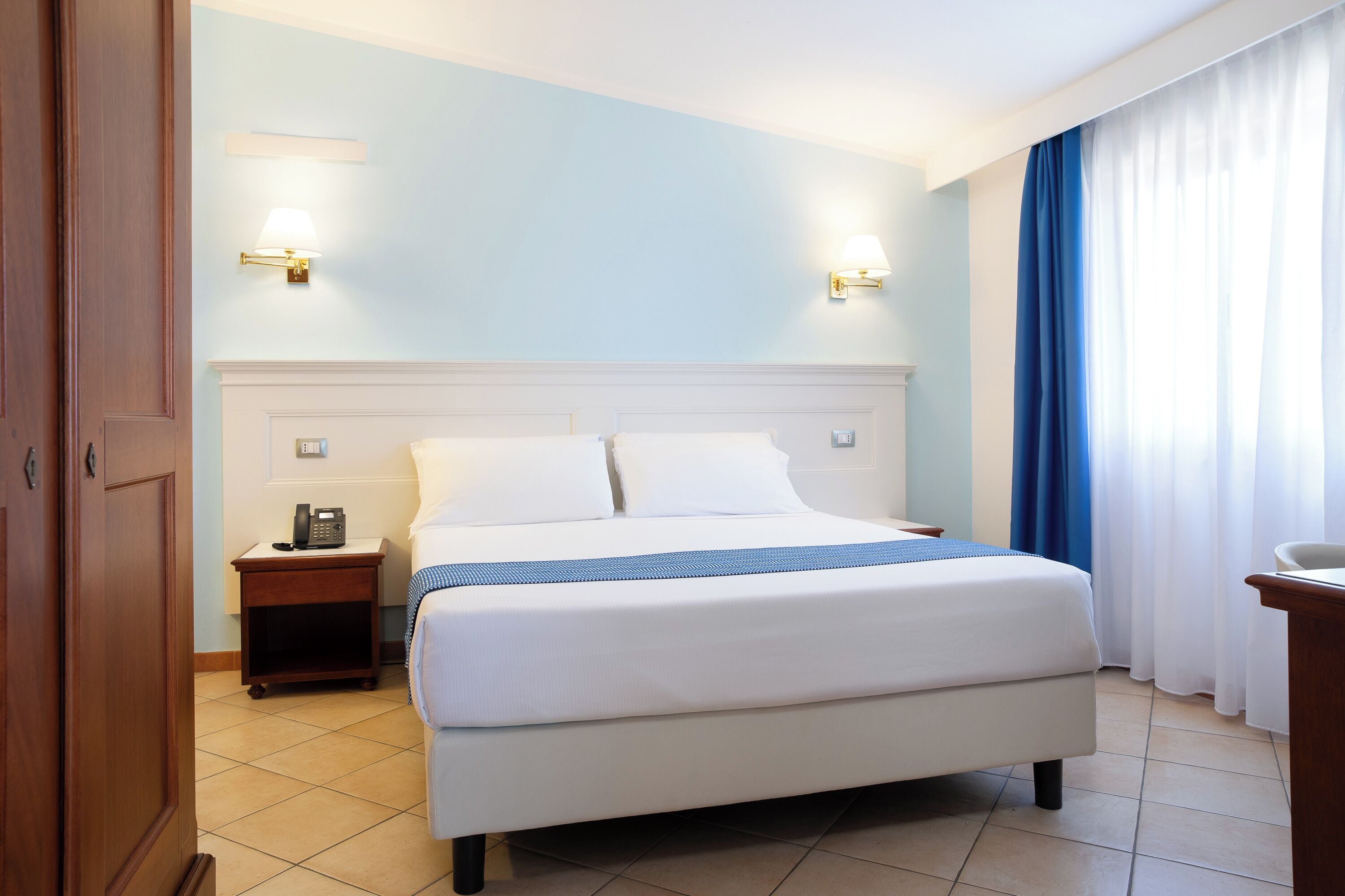 Une chambre d’hôtel avec un grand lit, deux tables de chevet, une petite table et une fenêtre avec des rideaux.