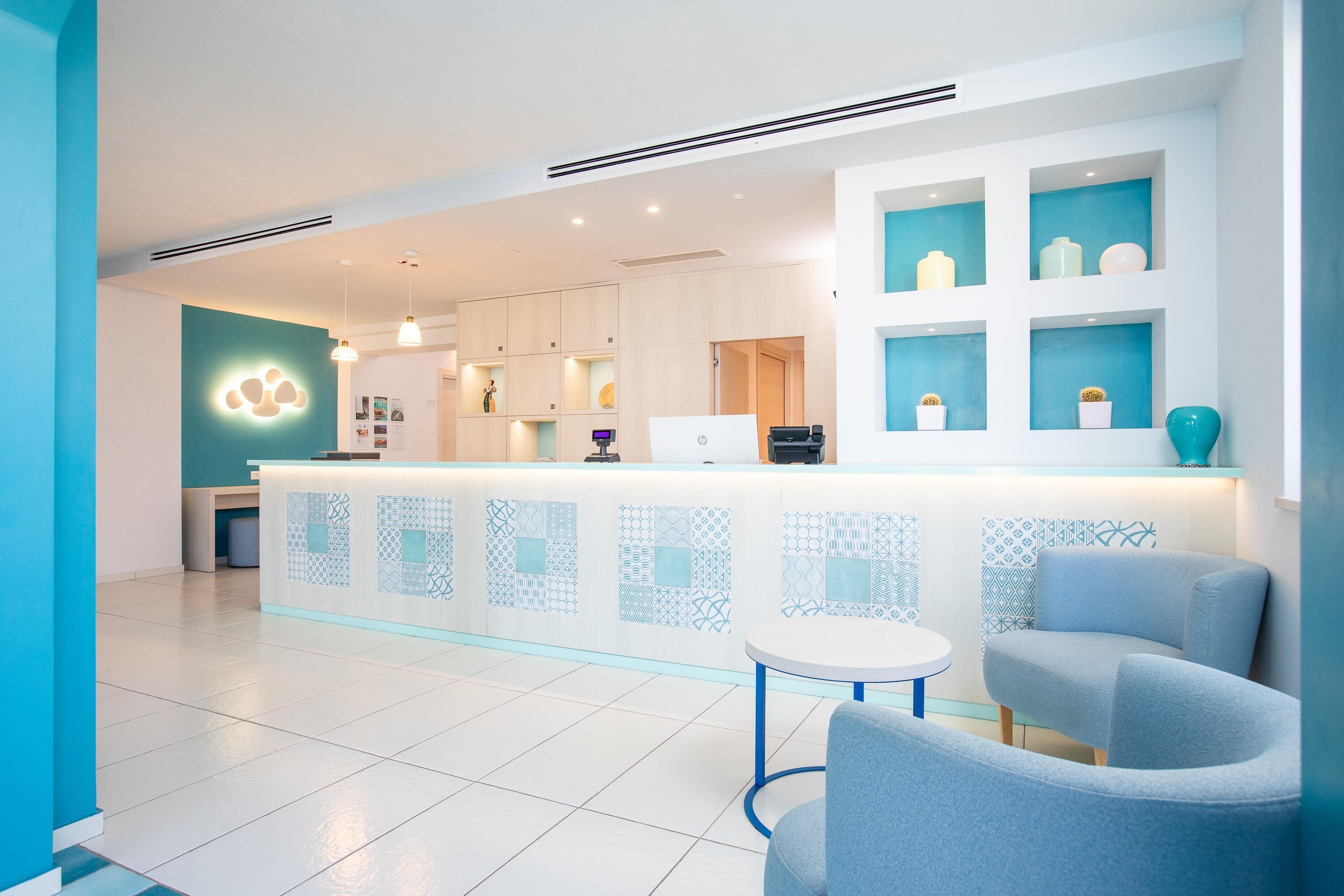 Una zona de recepción moderna con mostrador blanco, baldosas con diseño azul y un área de descanso con sillas grises.