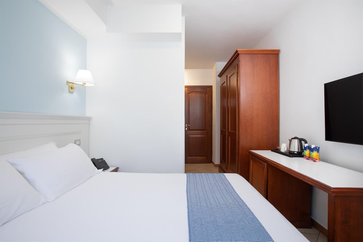 Camera d'albergo con un letto, una lampada da comodino, un armadio in legno, una TV a schermo piatto e un tavolino con bollitore e tazze.