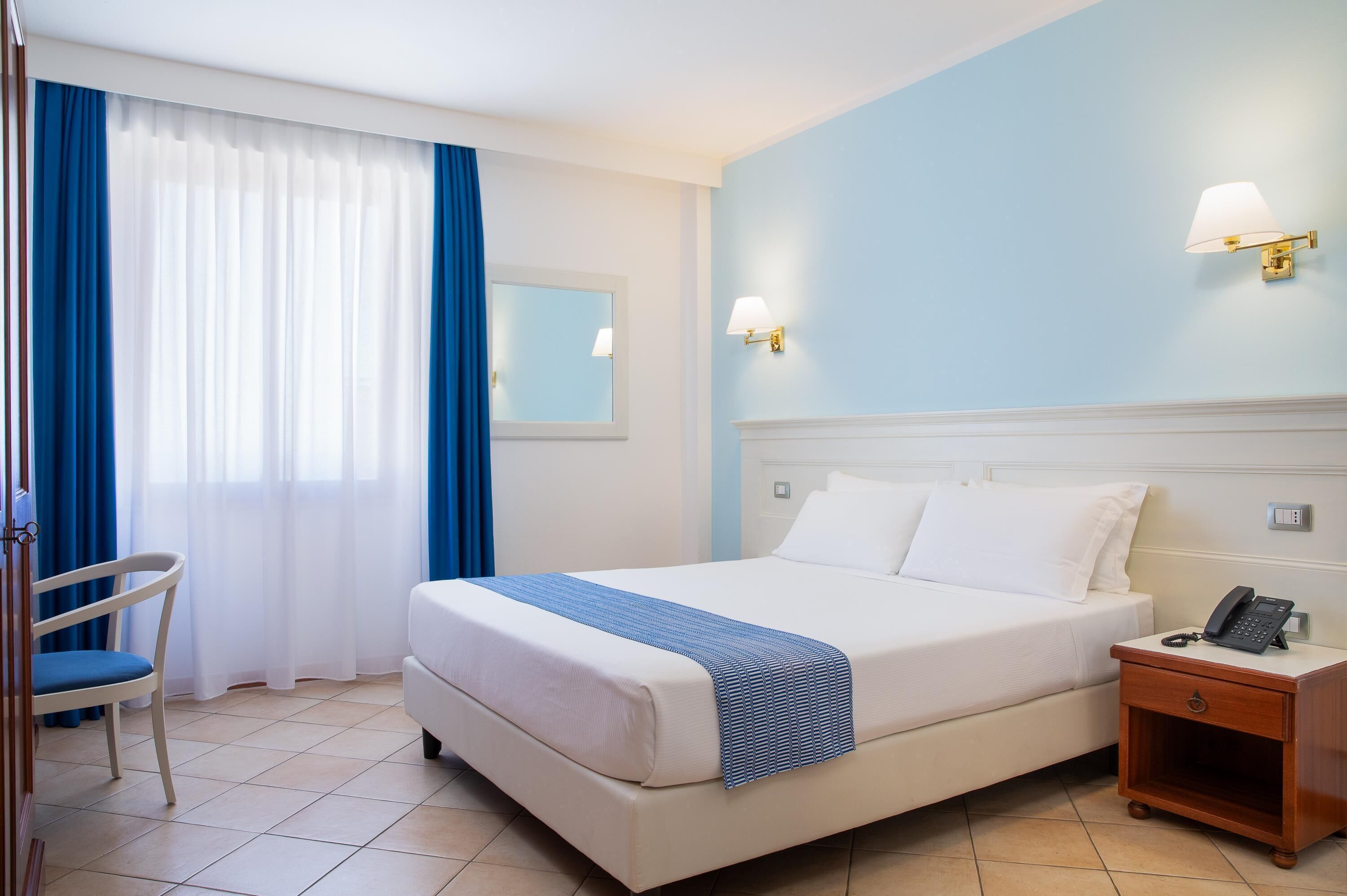 Una habitación de hotel con cama, una silla, una mesita de noche con teléfono y una ventana con cortinas.