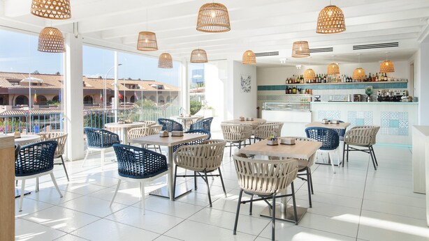 A spacious outdoor dining area with wicker chairs and tables, a bar with a tiled backsplash, and large windows offering views of buildings and a clear sky. - Hotel Blumarea, BW Signature Collection (Castelsardo)