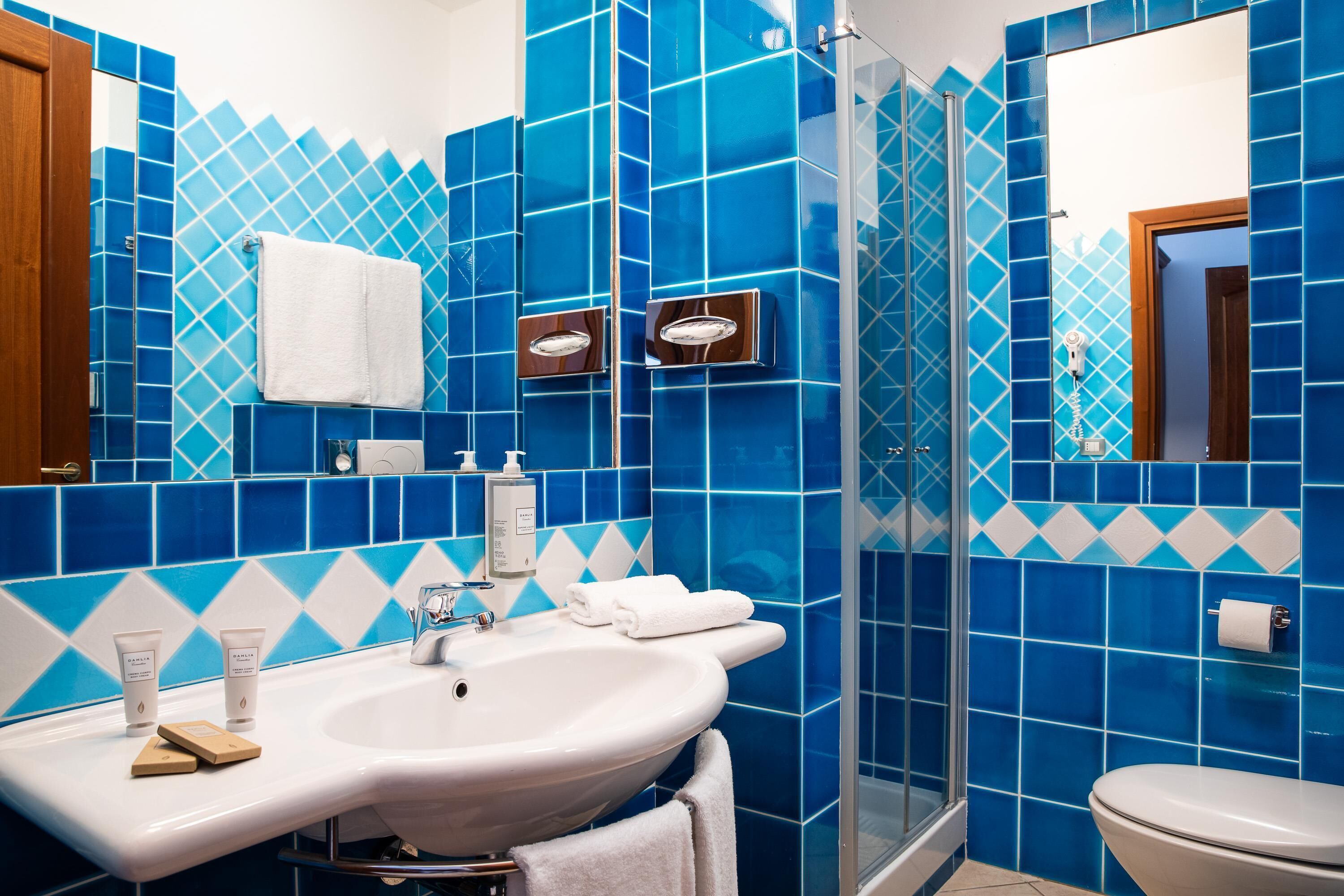Een badkamer met een wit wastafelmeubel, blauwe tegels, een glazen douchecabine en een toilet.